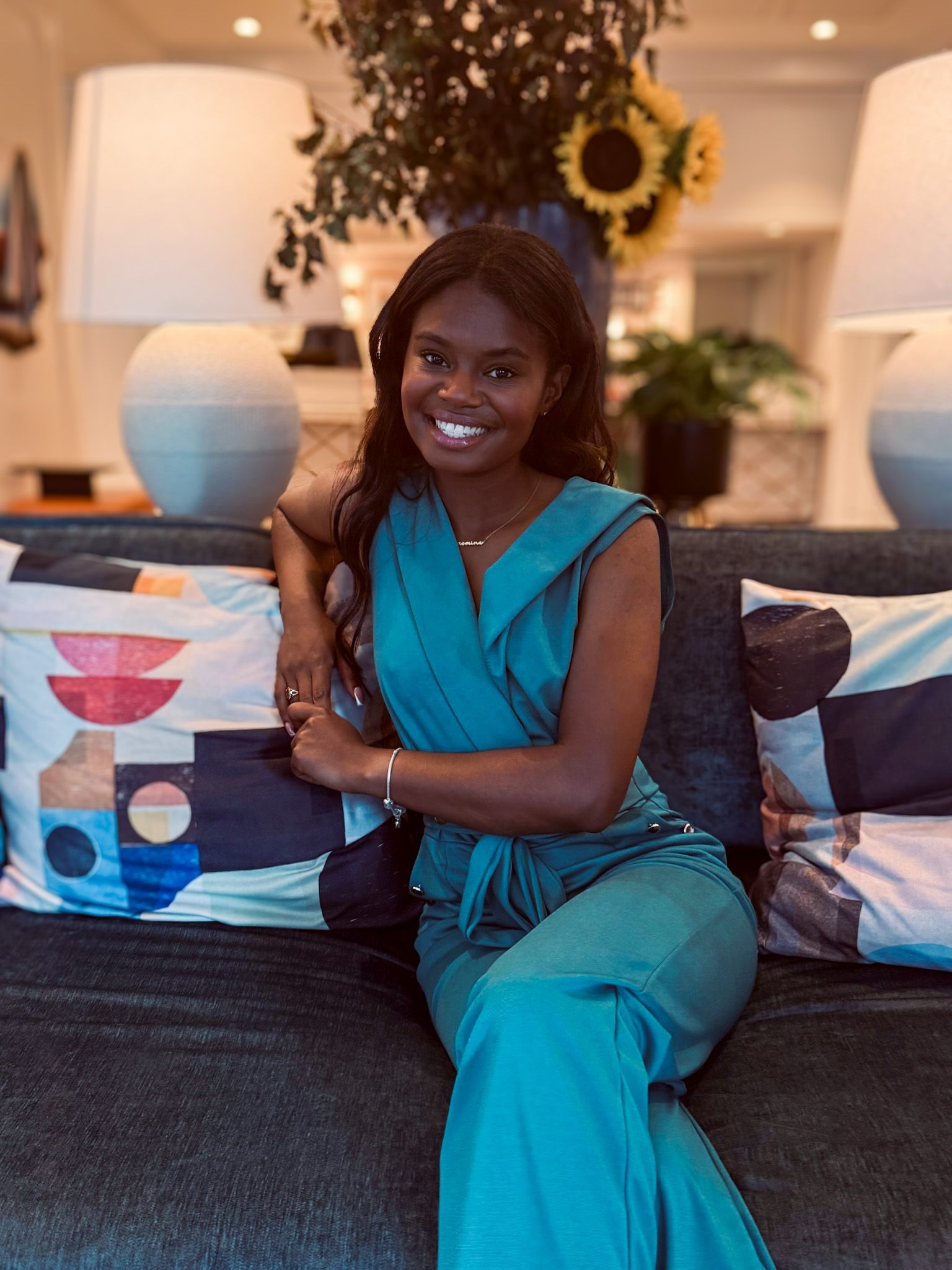 A woman is sitting on a couch in front of two lamps and a vase of flowers.
