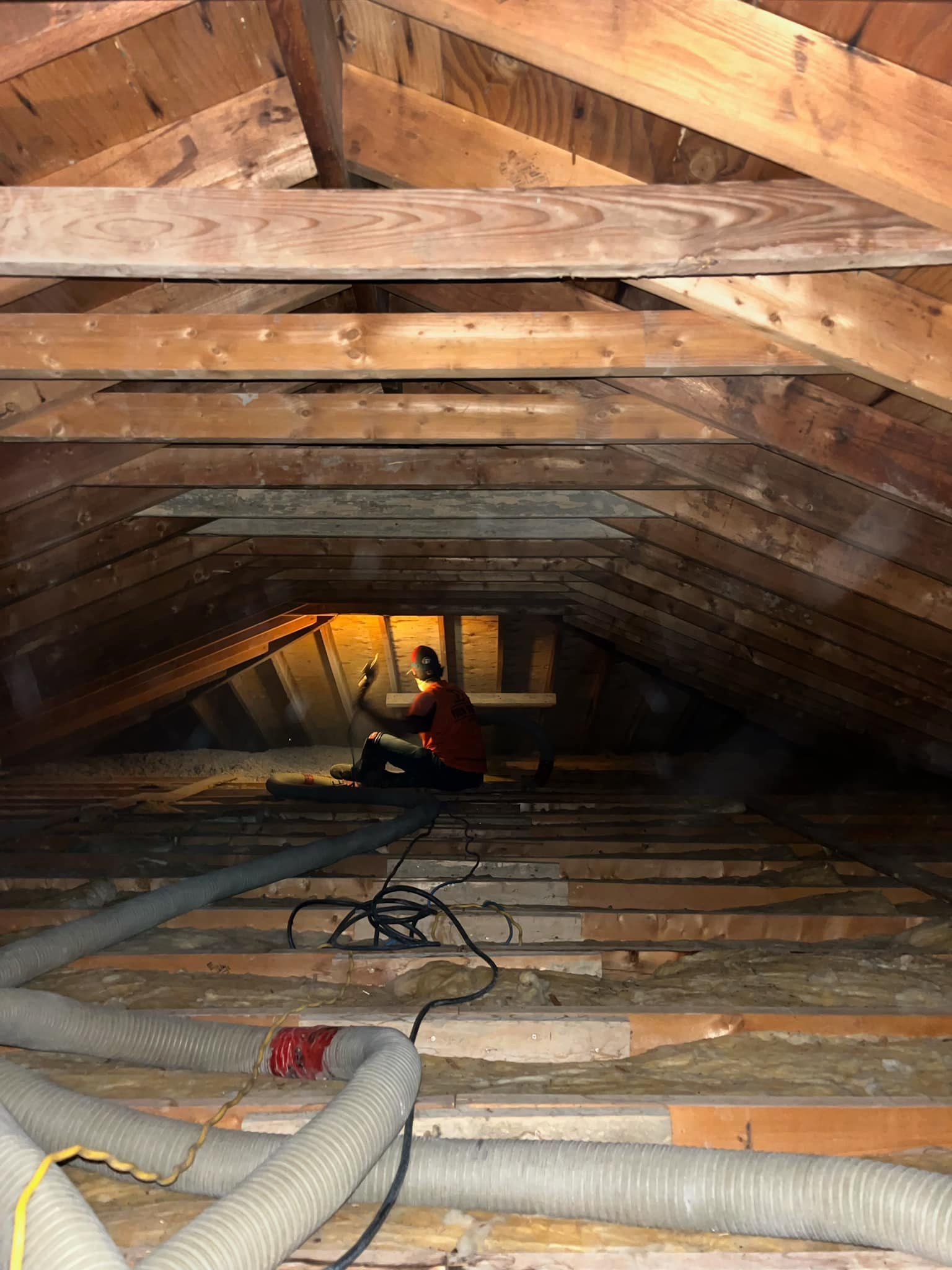 Person in an attic with insulation, working with a hose and wires. Wooden beams and rafters are visible.