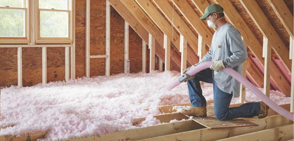 A worker insulates an attic with pink insulation, kneeling.