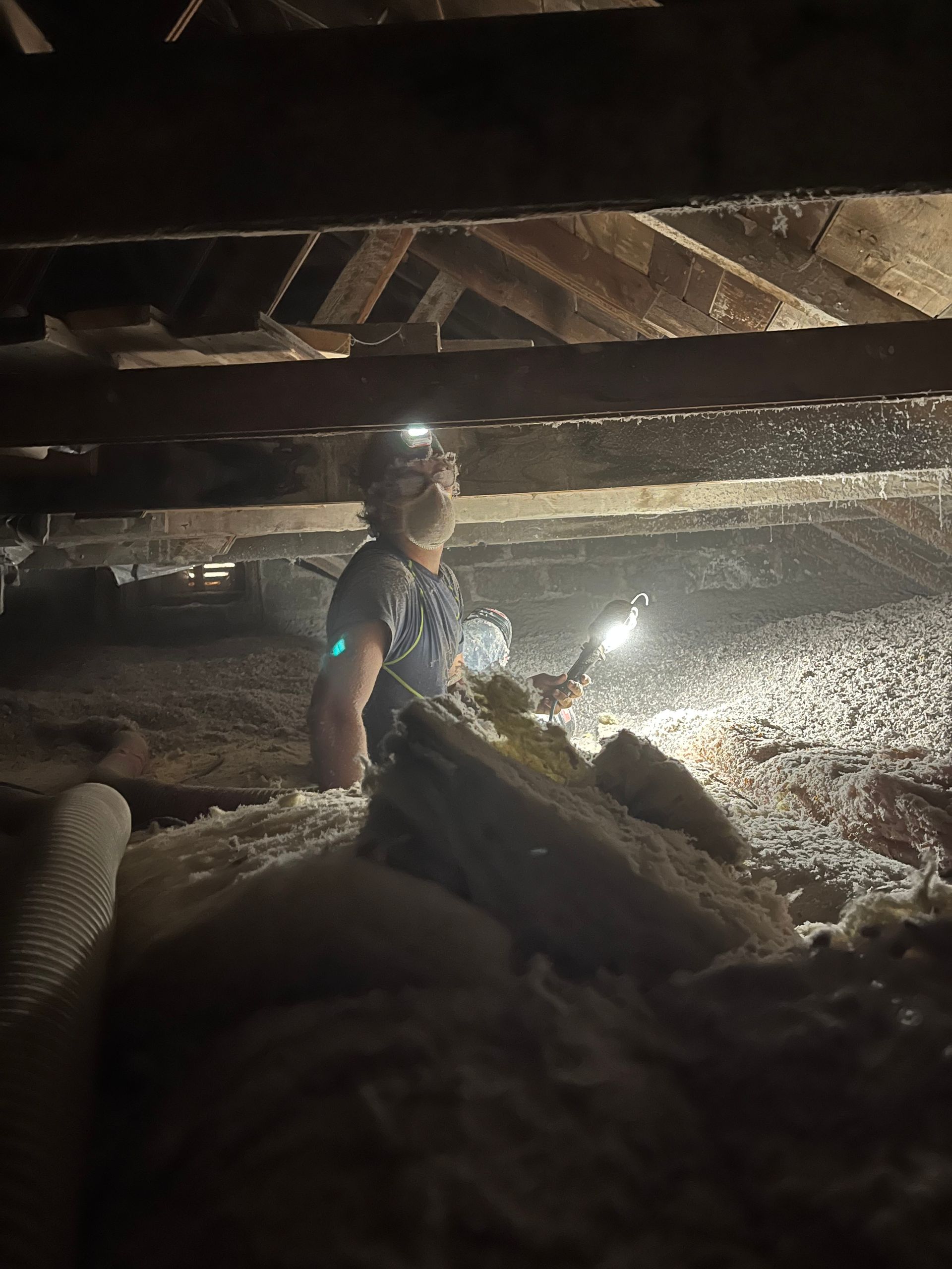 Person in a dark attic, wearing a headlamp and holding a flashlight, surrounded by insulation.