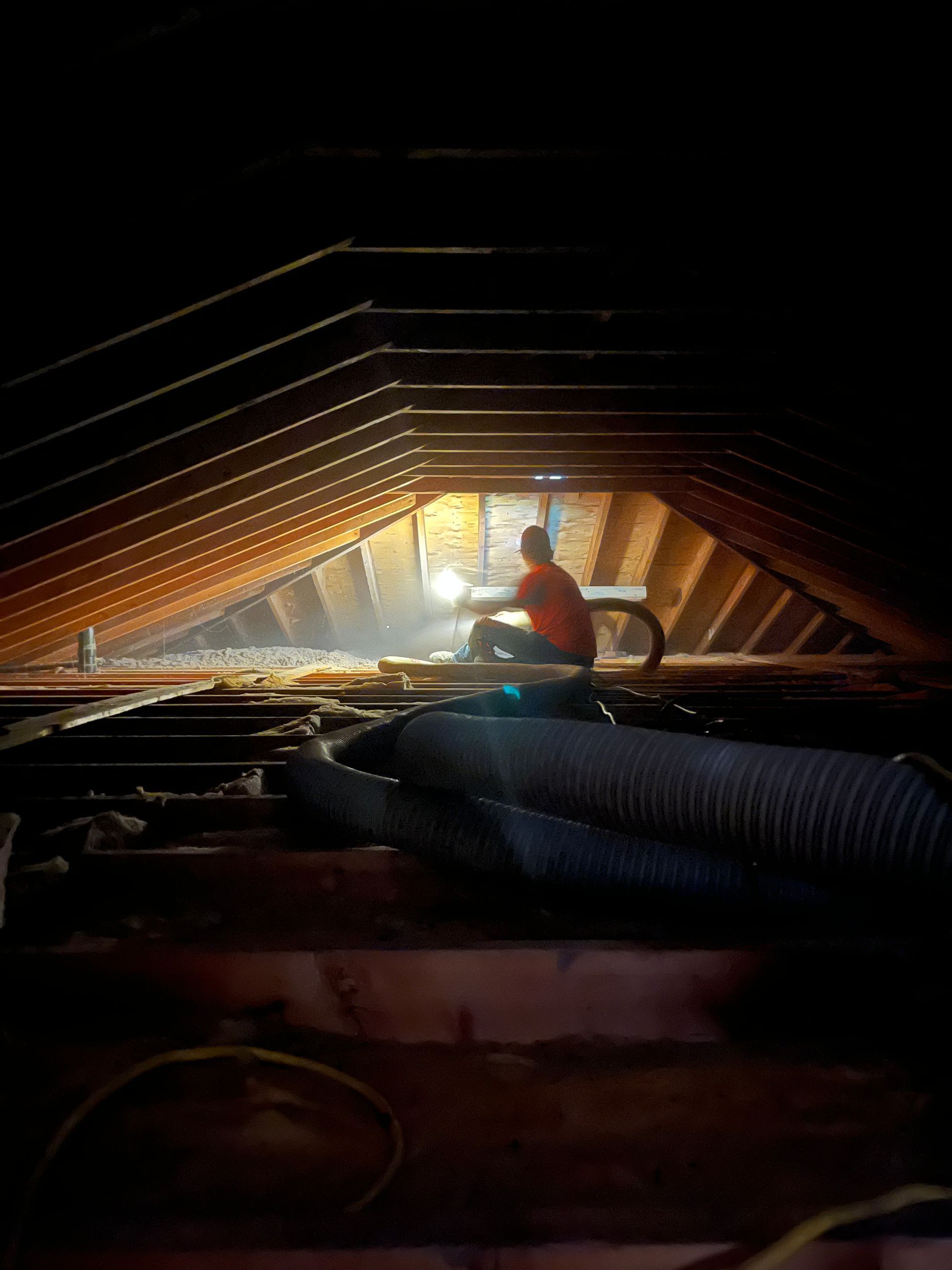 Person working in a dark attic with sunlight, surrounded by wood beams and insulation.