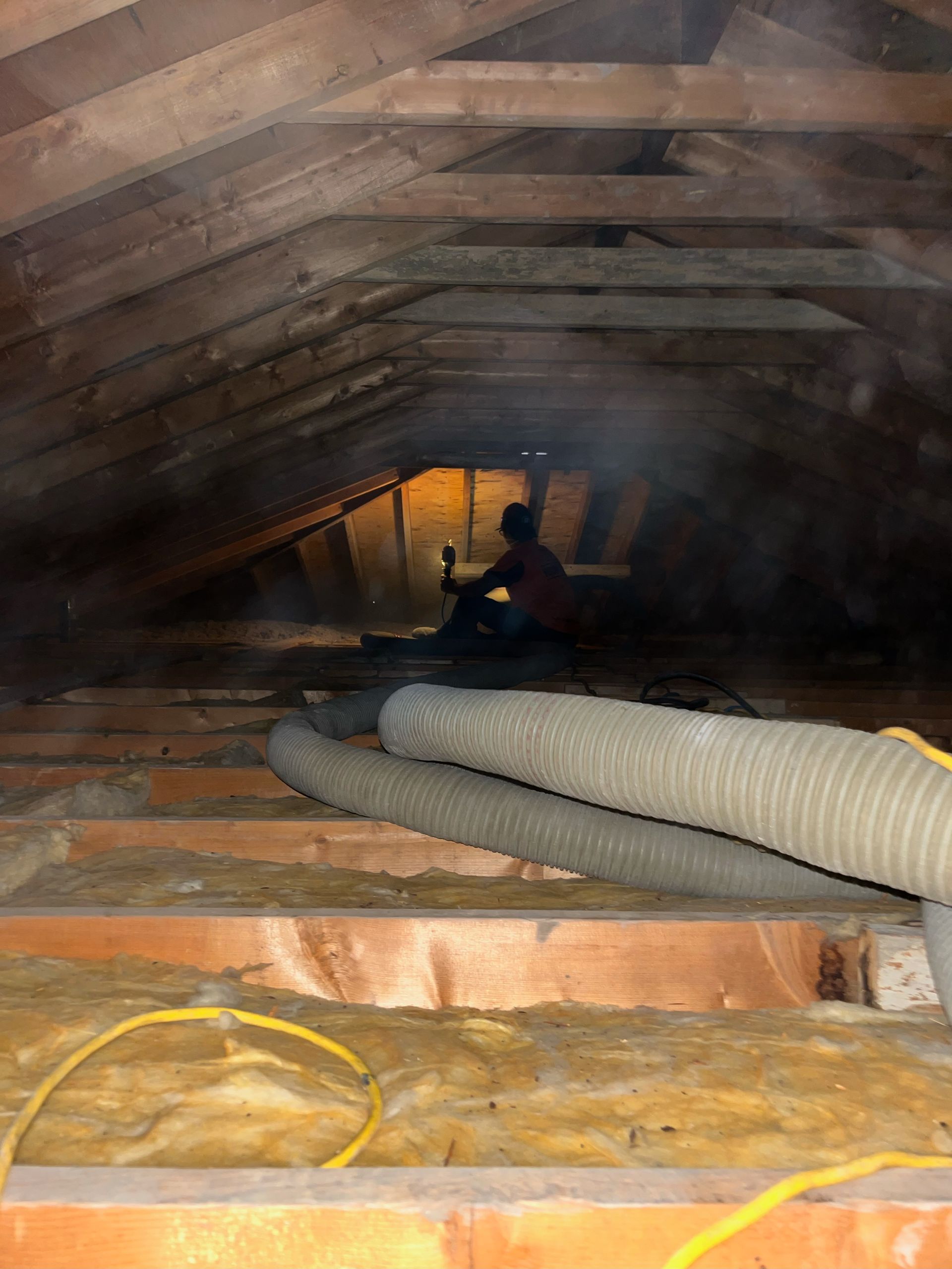 Person in an attic, holding a light. Insulation, ducting, and wood beams are visible.