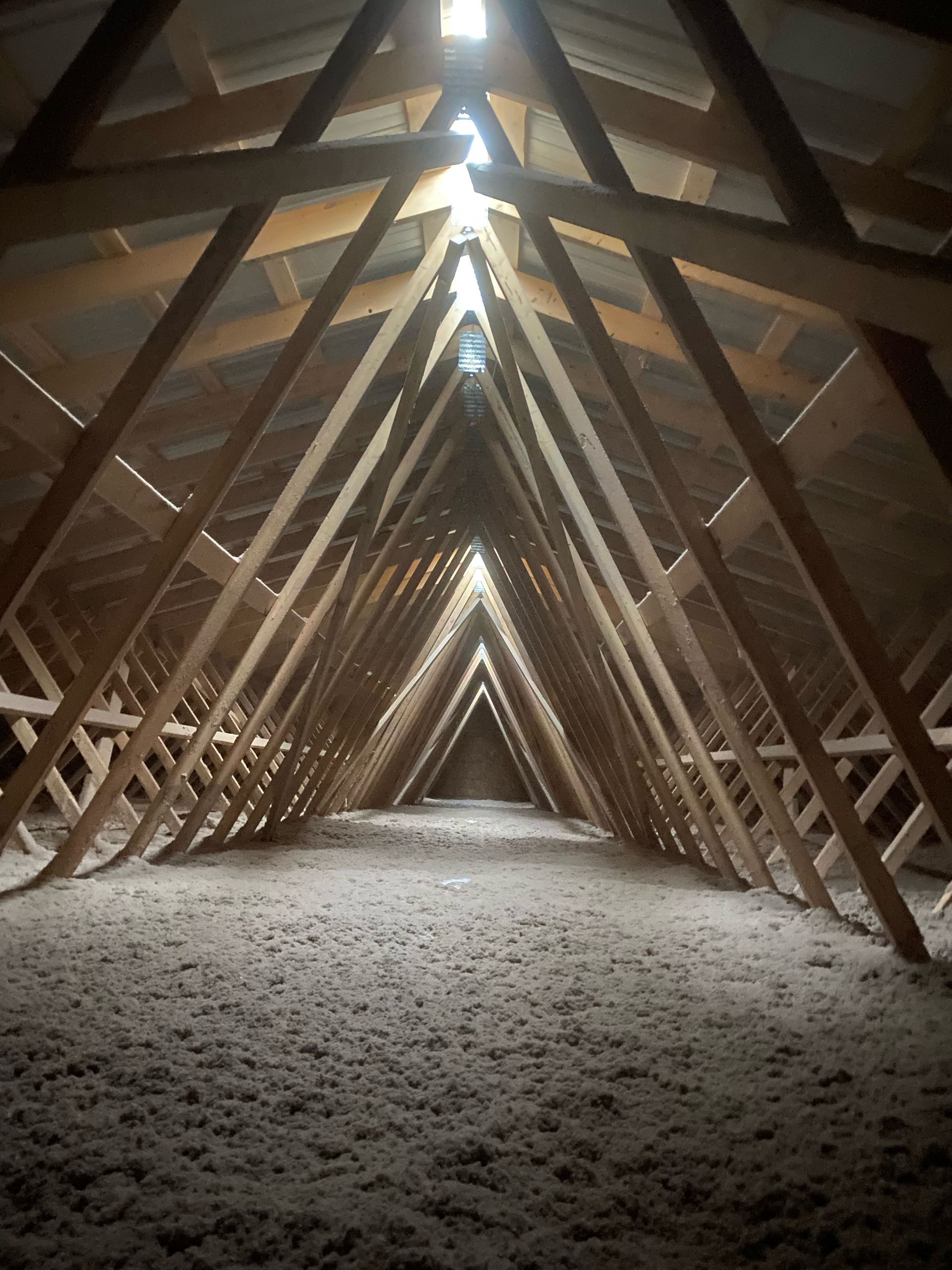 Interior view of an attic, with wooden rafters and insulation, perspective leading to light at the apex.
