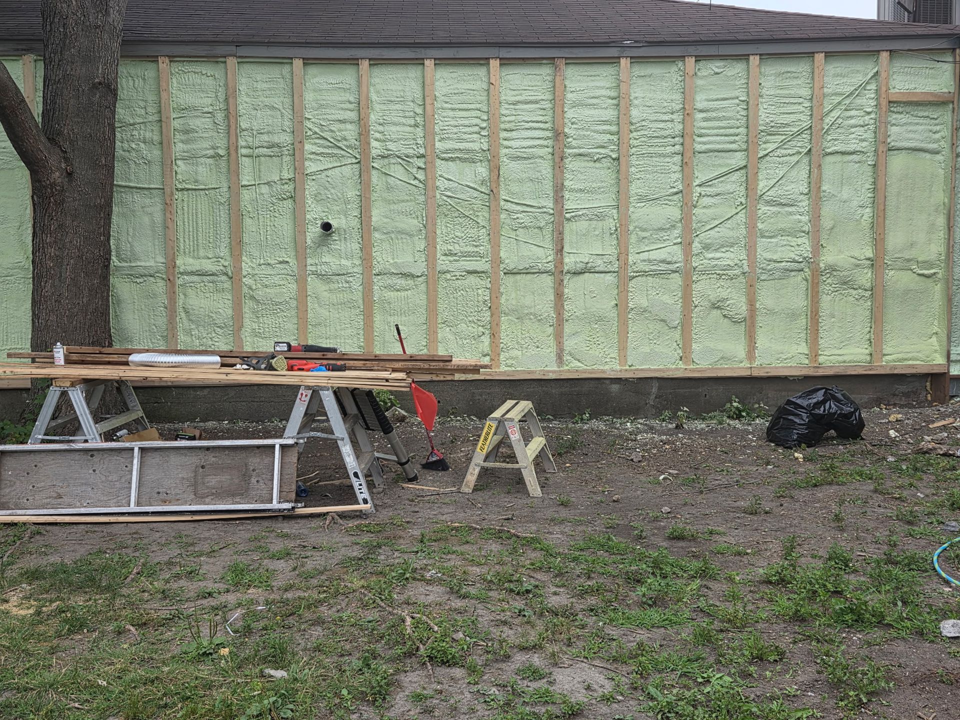 Exterior wall under construction with insulation and framing. Tools, sawhorses, and a tree are in the yard.