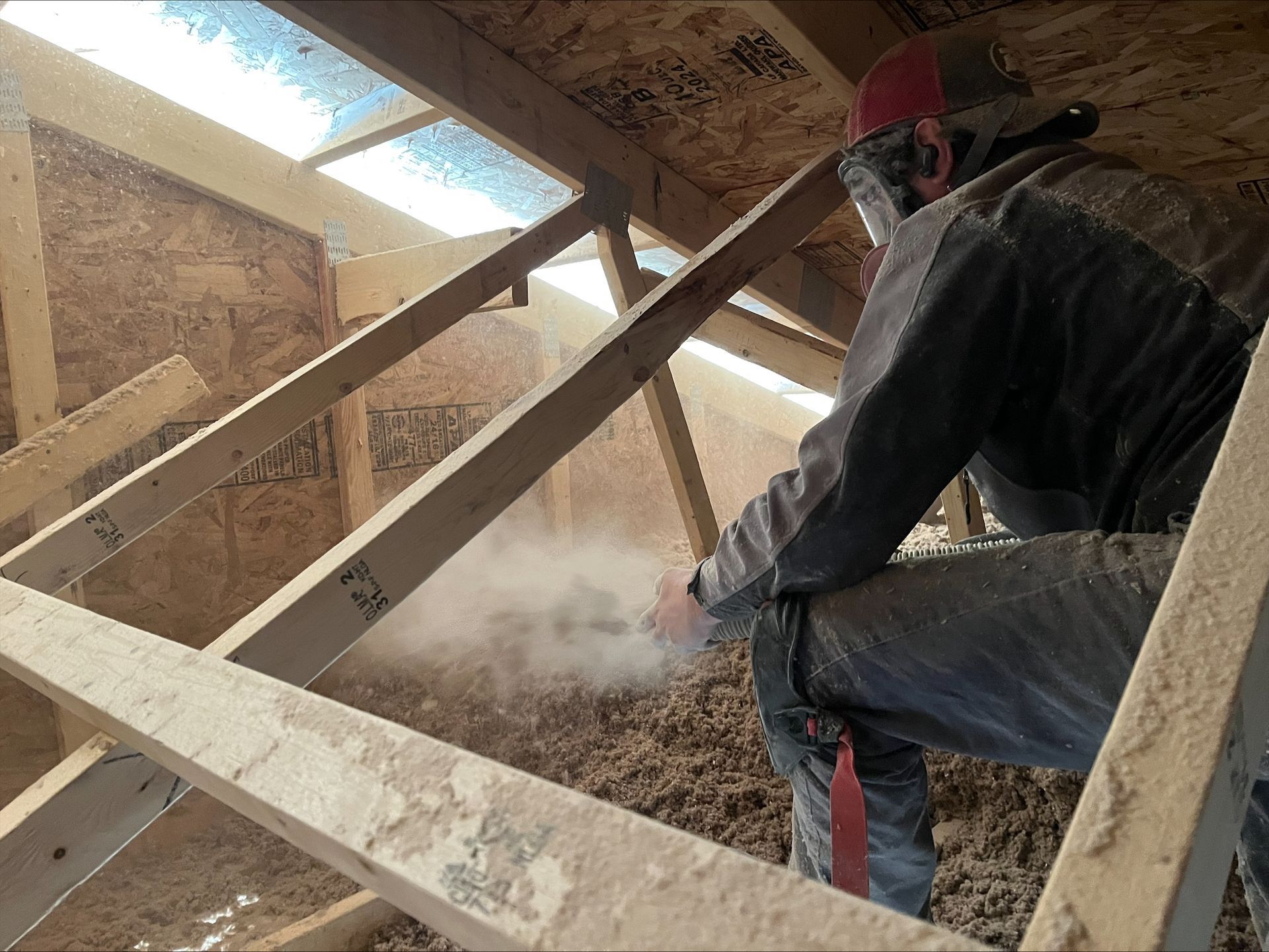 Person in protective gear blowing insulation into an attic, dusty environment.
