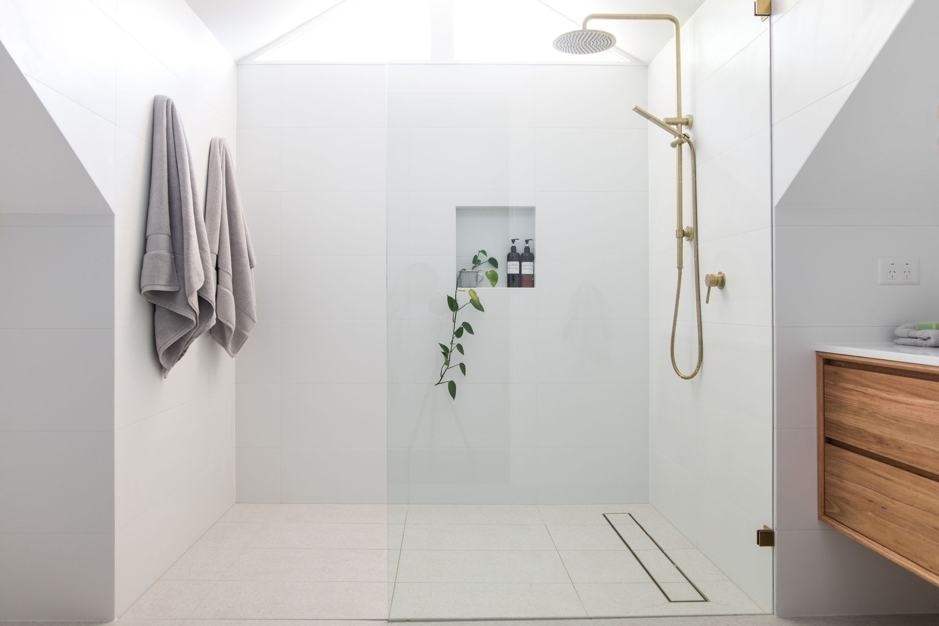 Modern white-tiled attic shower with gold tap, timber vanity, and soft grey towels