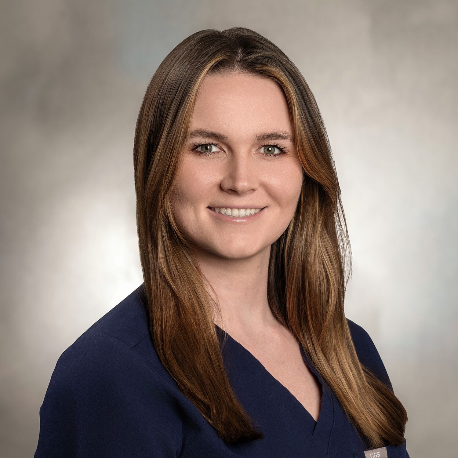 Woman with long brown hair, smiling, wearing a dark blue top, against a blurred background.