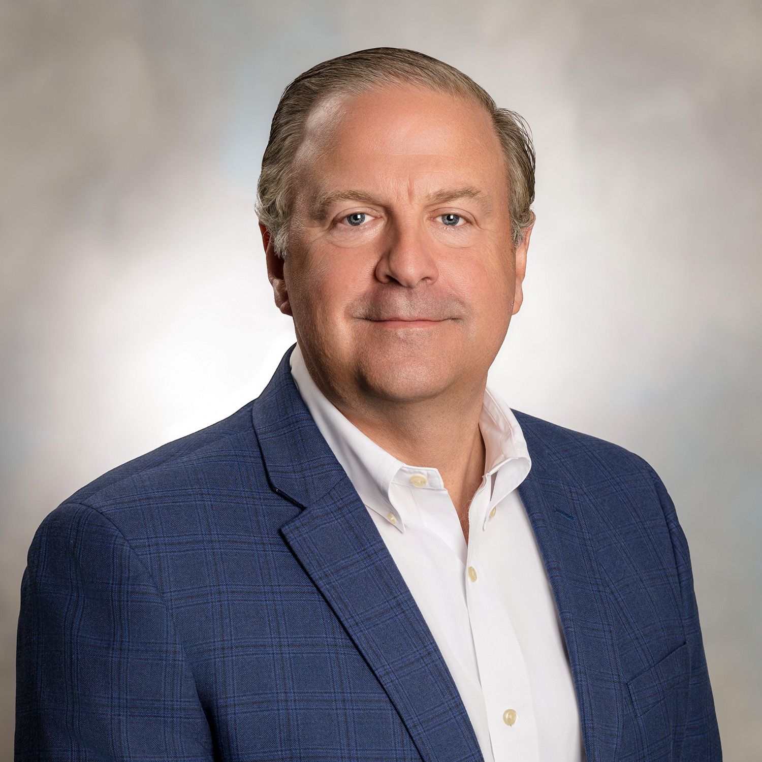 Man in blue blazer and open-collared white shirt smiles, against a blurred gray-blue backdrop.