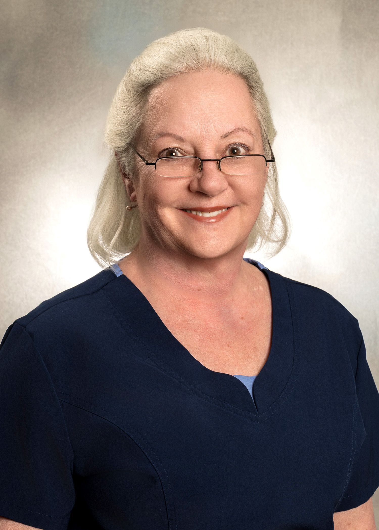 Woman with glasses wearing blue scrubs smiles.