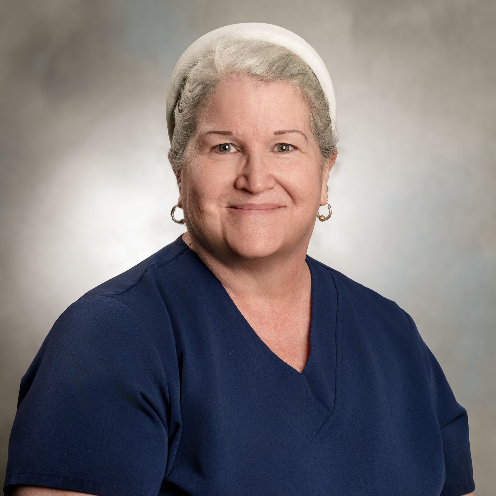 Woman wearing a blue scrub top and white head covering, smiling, against a gray background.