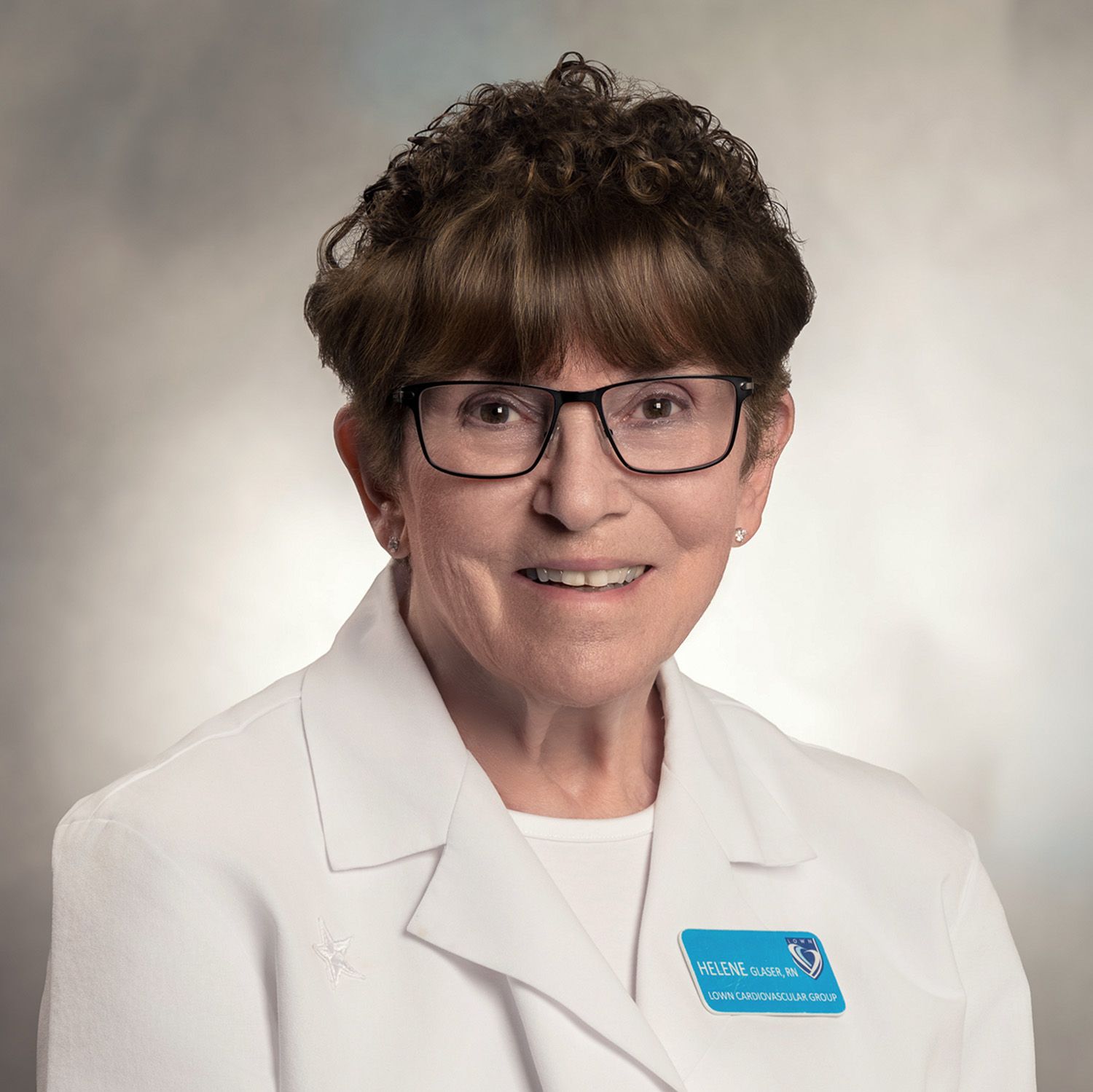 Woman wearing glasses, a white coat, and a name tag smiles at the camera.