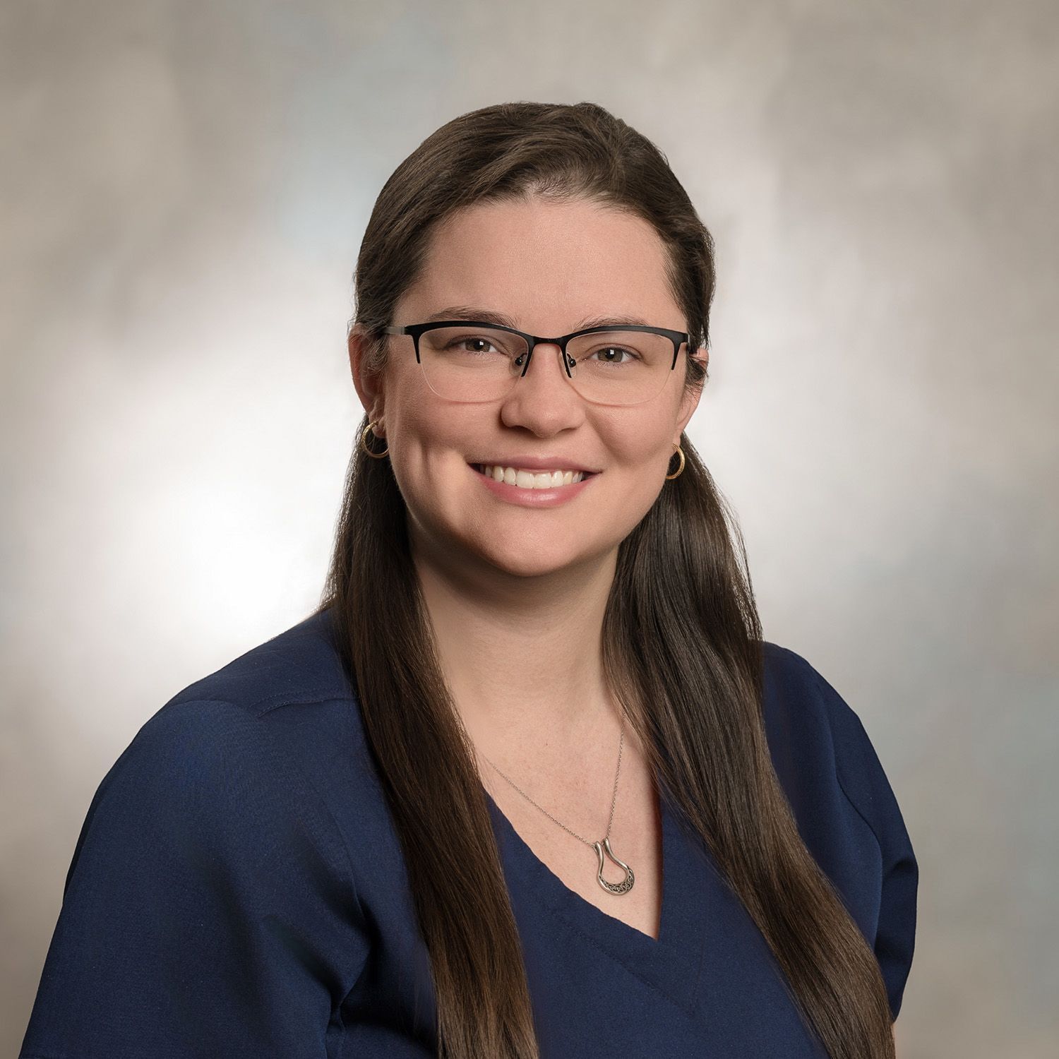 Woman with glasses and long brown hair smiles, wearing a blue top, against a neutral background.