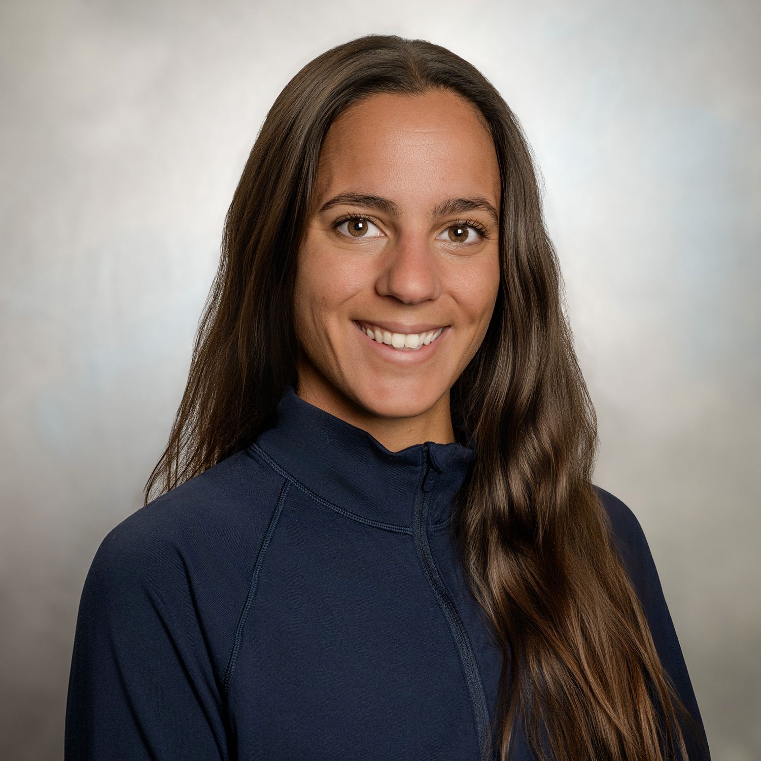 Woman with long brown hair smiles, wearing a blue zip-up jacket.