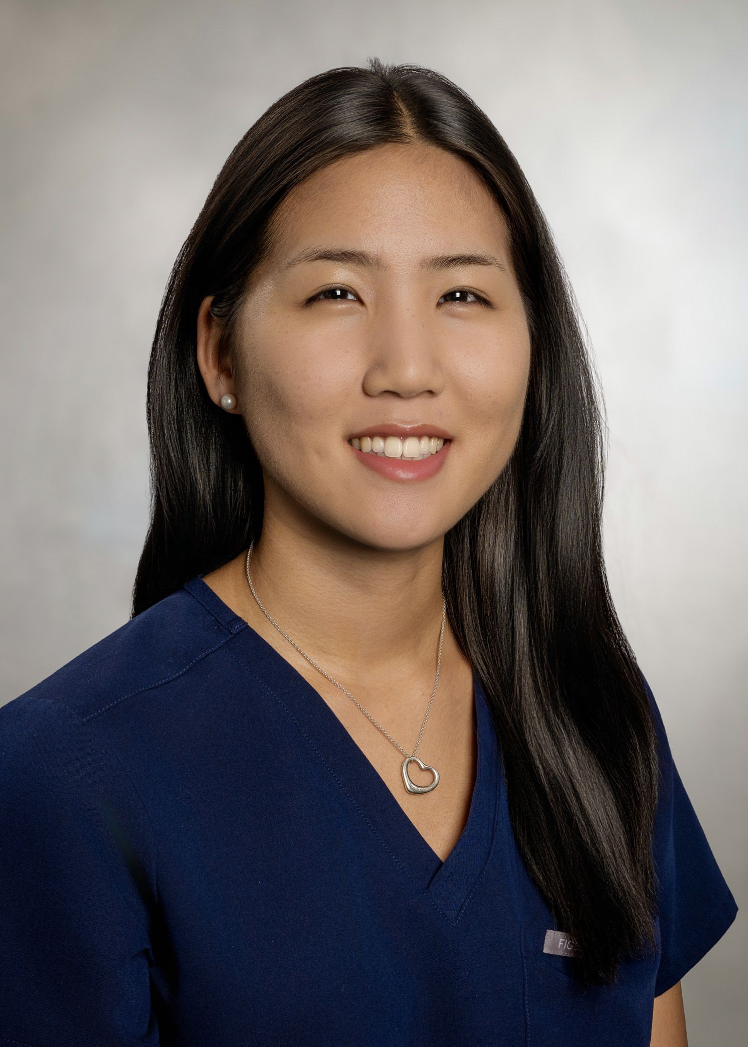 Woman with long black hair smiles, wearing a blue top and a necklace against a gray background.