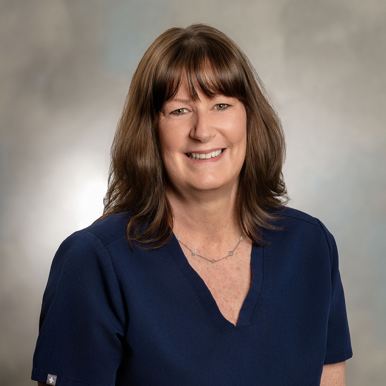 Woman with brown hair and bangs smiles, wearing a blue top, against a gray gradient background.