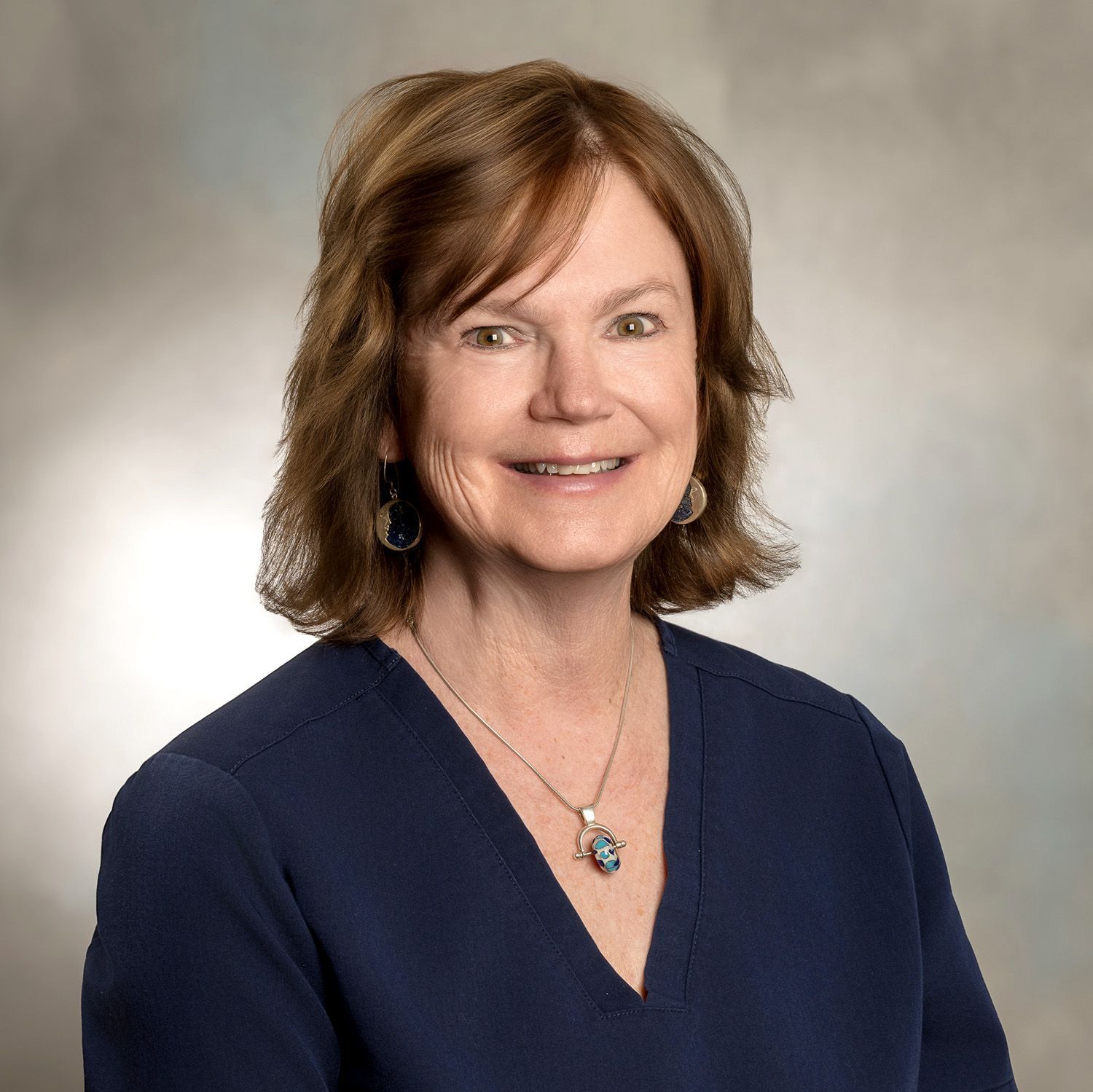 Woman with auburn hair in a blue top smiles, wearing a necklace. Studio setting with a blurred background.
