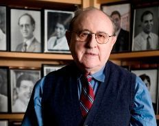 A man wearing glasses and a tie is standing in front of a wall of pictures.