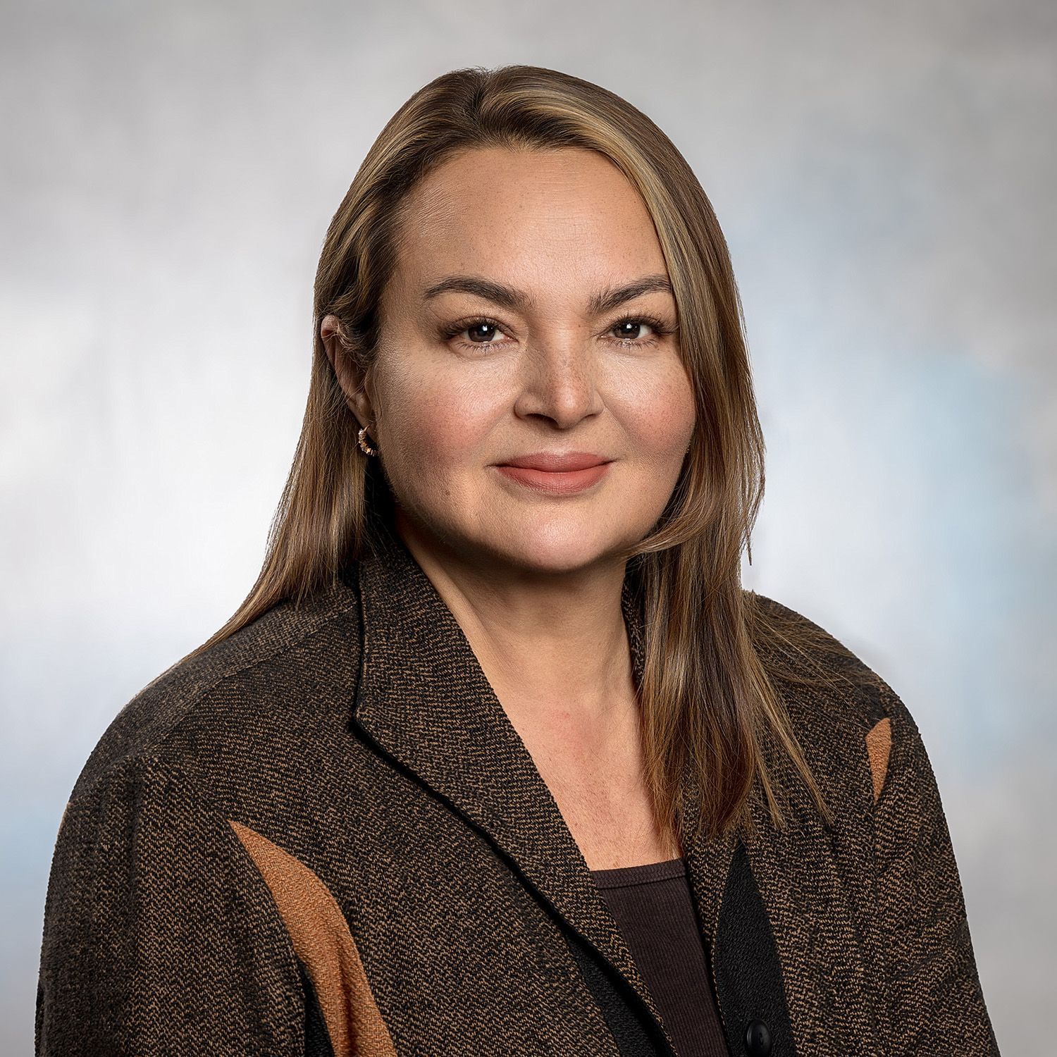 Woman in a brown jacket smiles slightly, against a blurred blue and white background.