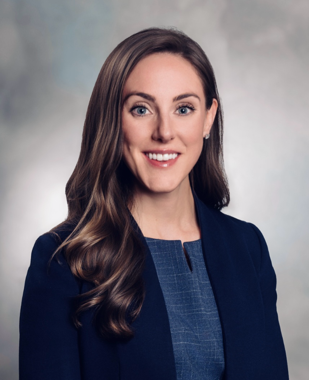 Woman with long brown hair, wearing a navy blazer and blue textured top, smiling.