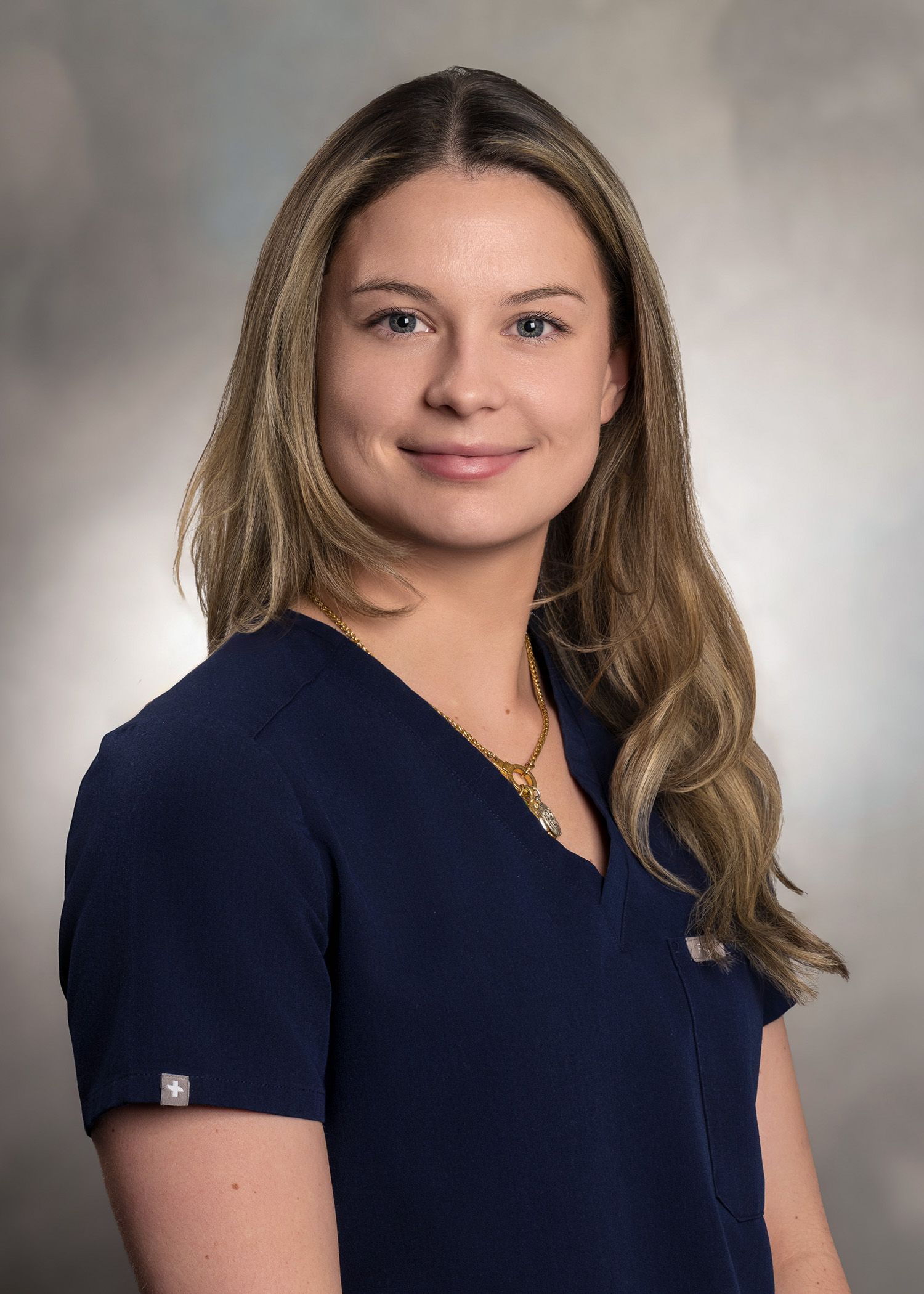 Woman with long, light-brown hair wearing a dark blue v-neck scrub top, smiling slightly, against a gray gradient backdrop.