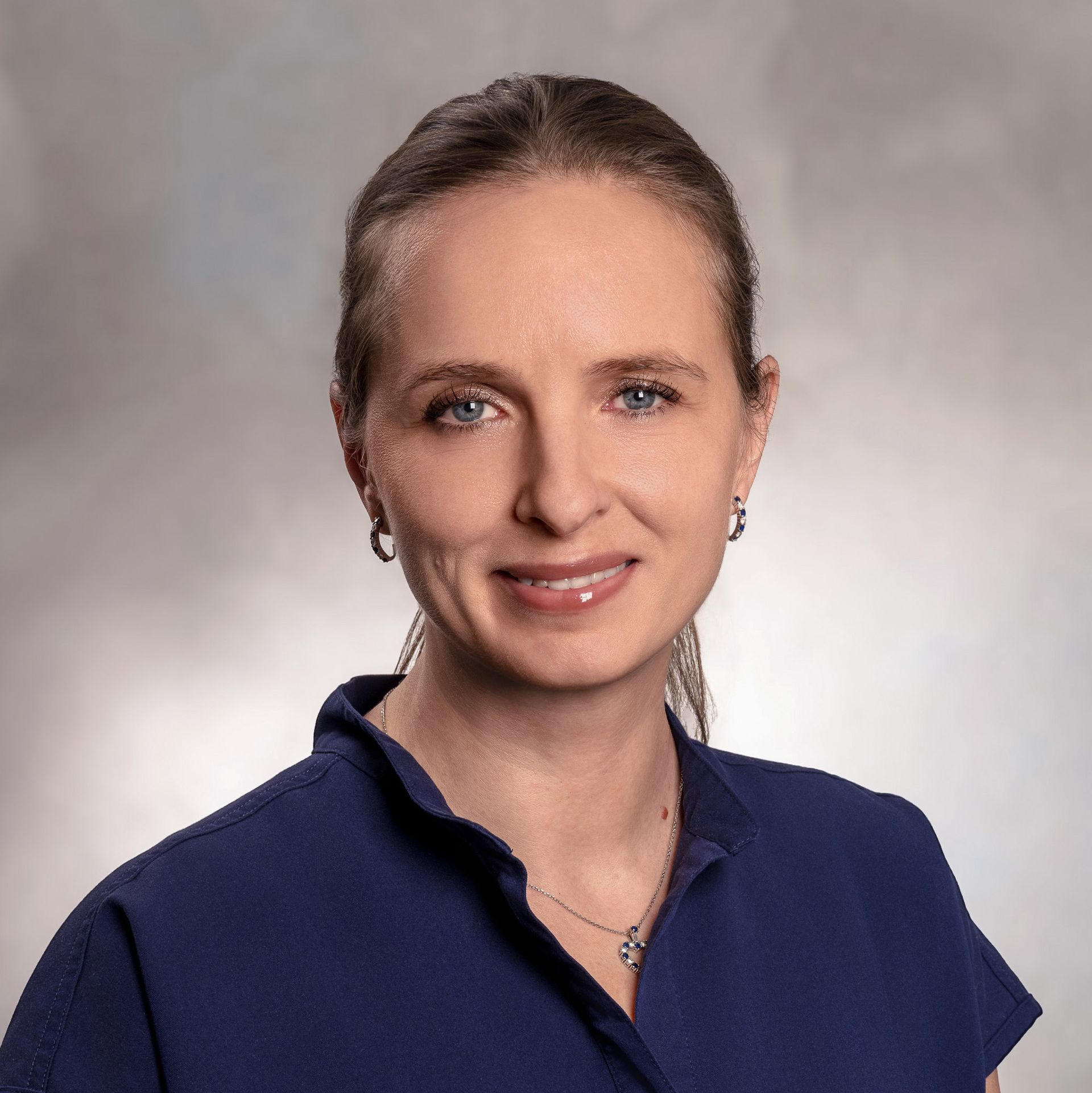 Woman with light skin, blue eyes, and pulled-back hair wearing a dark blue top smiles, against a blurred backdrop.