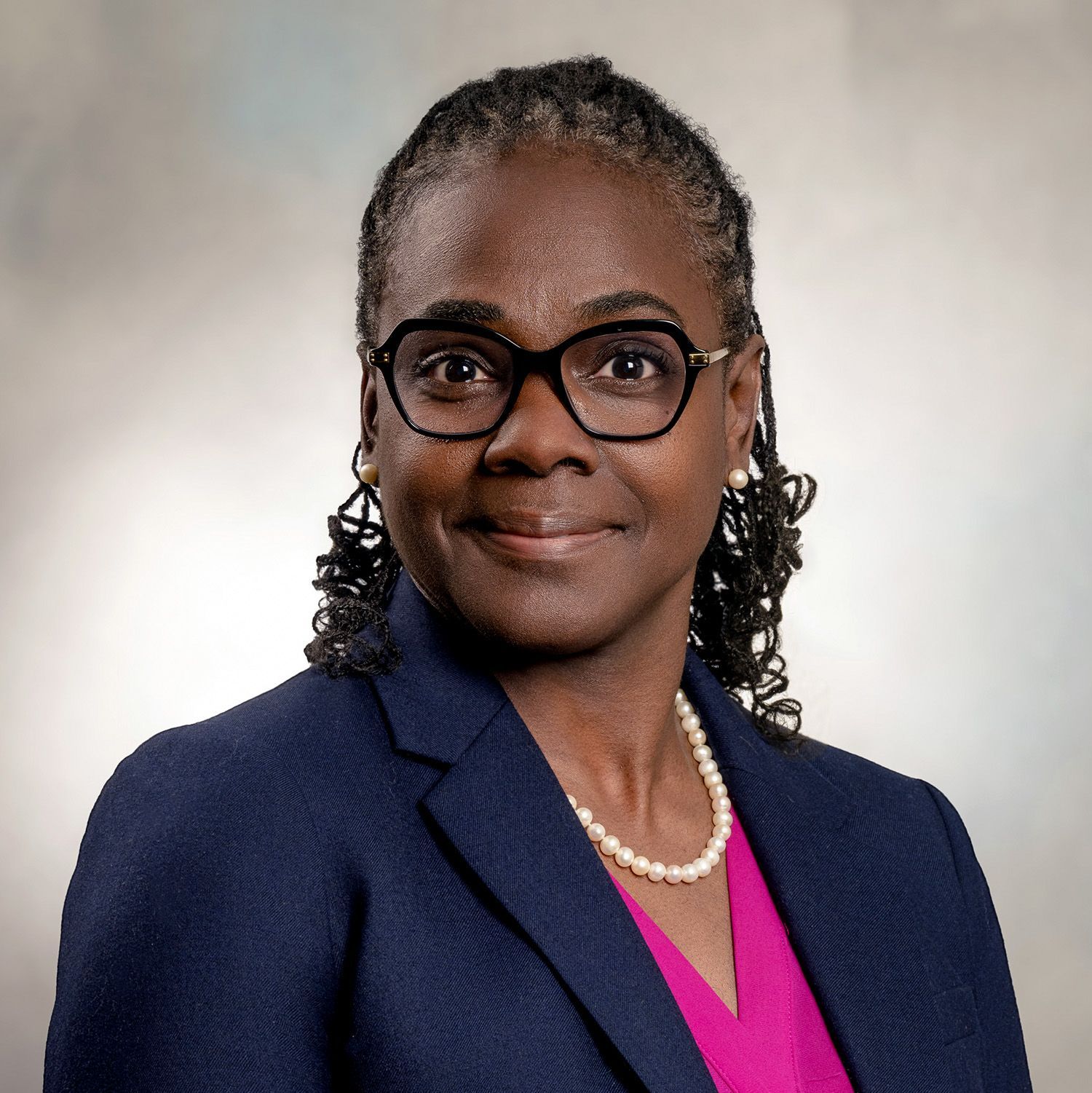 Woman with glasses, wearing a dark blue blazer and pink top, pearl necklace, smiling.