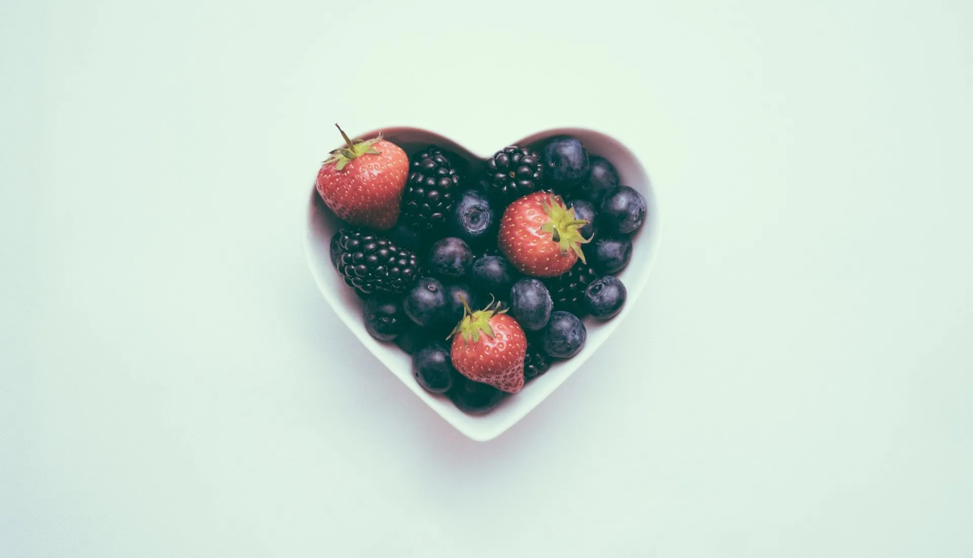 Heart-shaped bowl filled with blueberries, strawberries, and blackberries on a white surface.