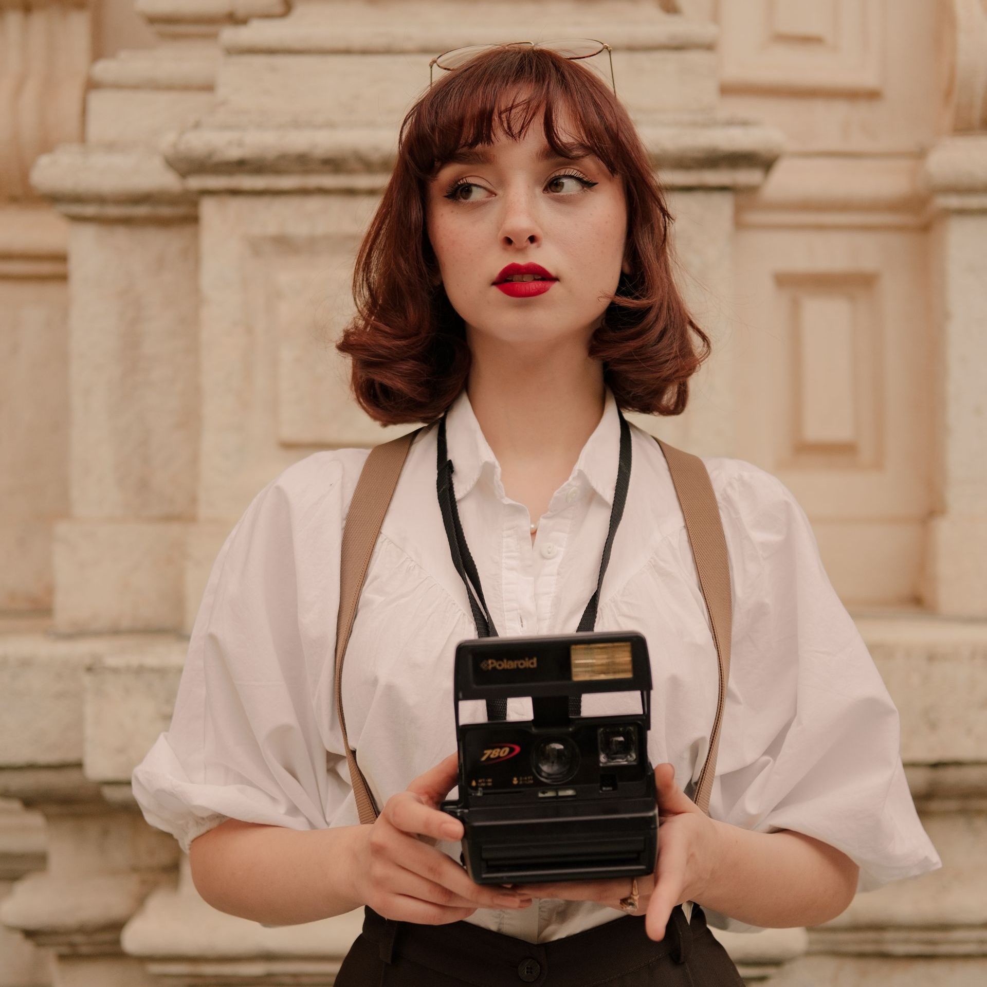 Vrouw met rood haar voor een oud gebouw. Ze draagt een witte blouse en heeft een vintage fotocamera.