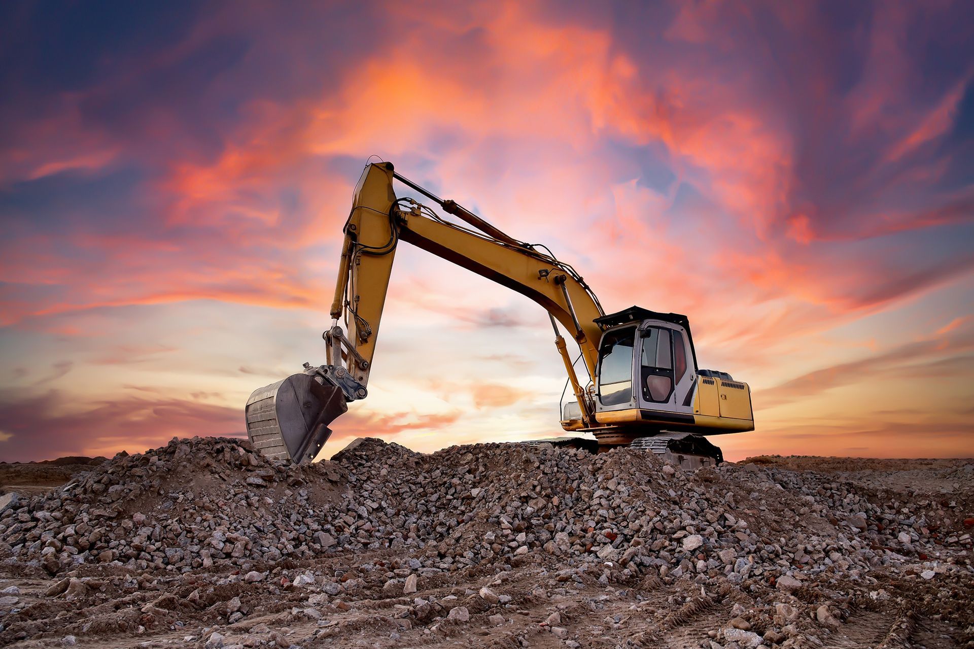 A yellow excavator is digging a pile of dirt in a field at sunset.