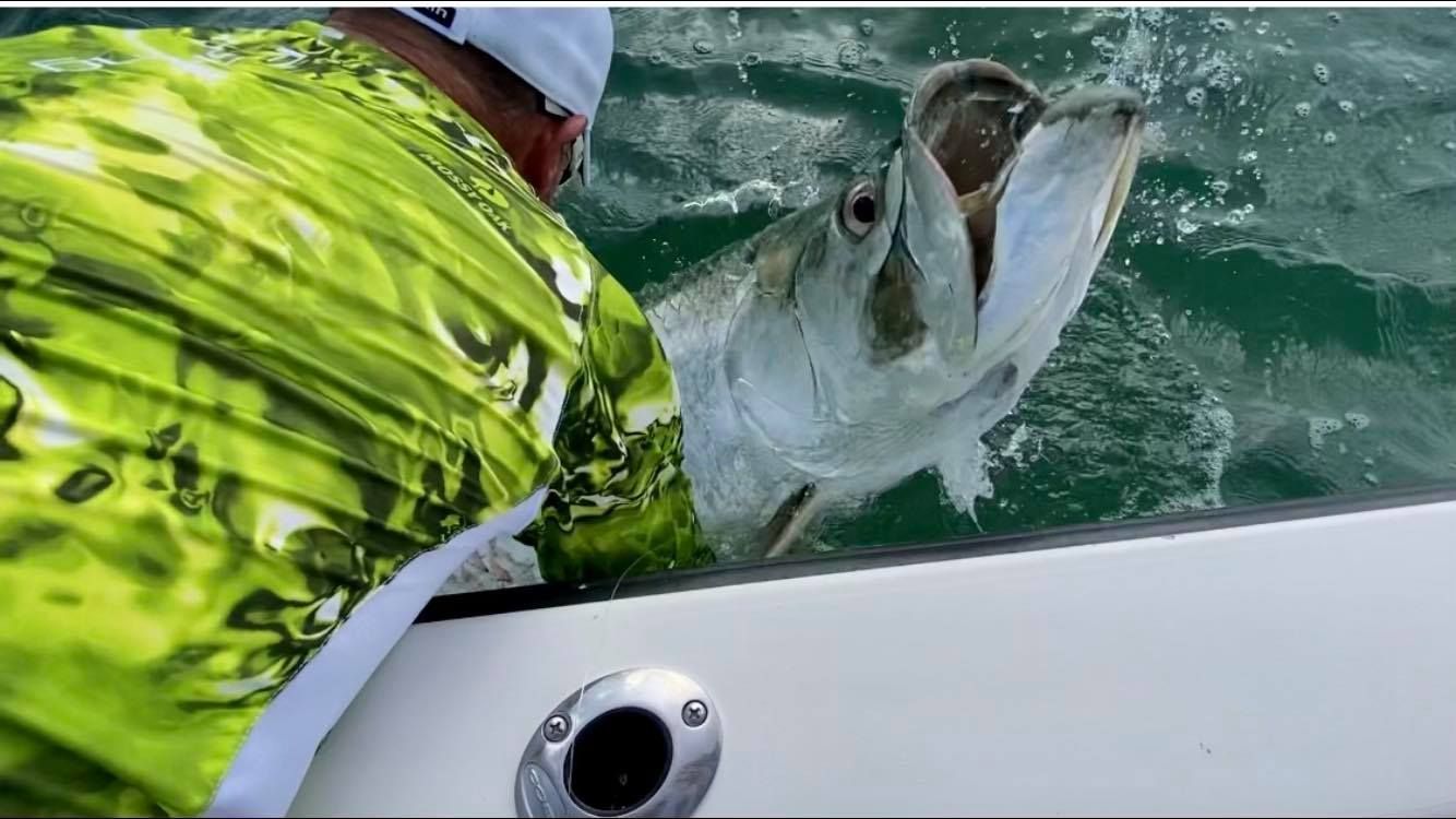 A man is fishing on a boat with a fish in his mouth.