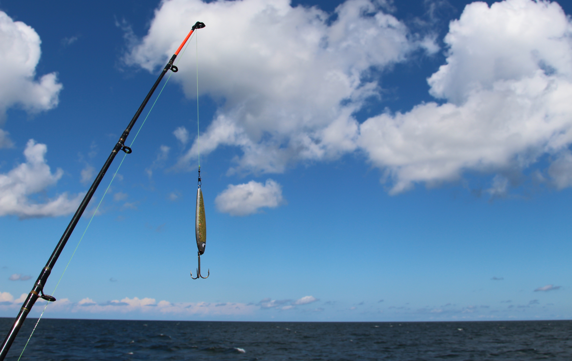 Fishing rod with lure hanging above ocean under a blue sky with clouds.