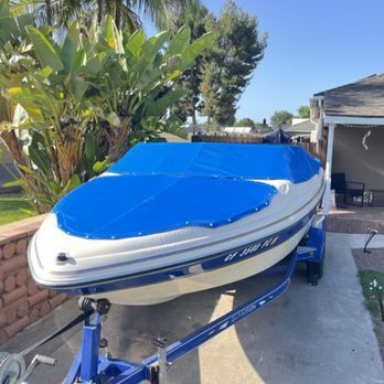 A white and blue boat on a trailer, covered with a blue tarp, parked in front of a house on a sunny day.