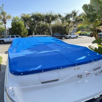 A white boat with a blue cover is parked in a driveway. Trees and houses are in the background.