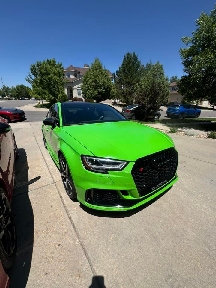 Bright green Audi RS3 parked on a sunny driveway. Black grille and roof, other cars in background.