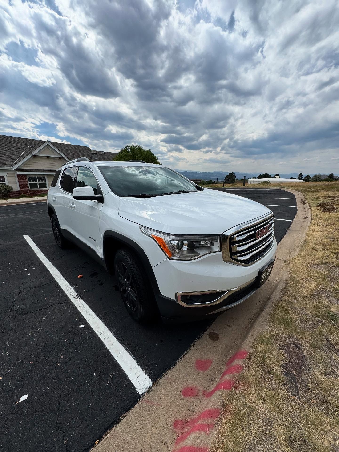 White GMC Acadia parked on asphalt next to curb and grass, under cloudy sky.