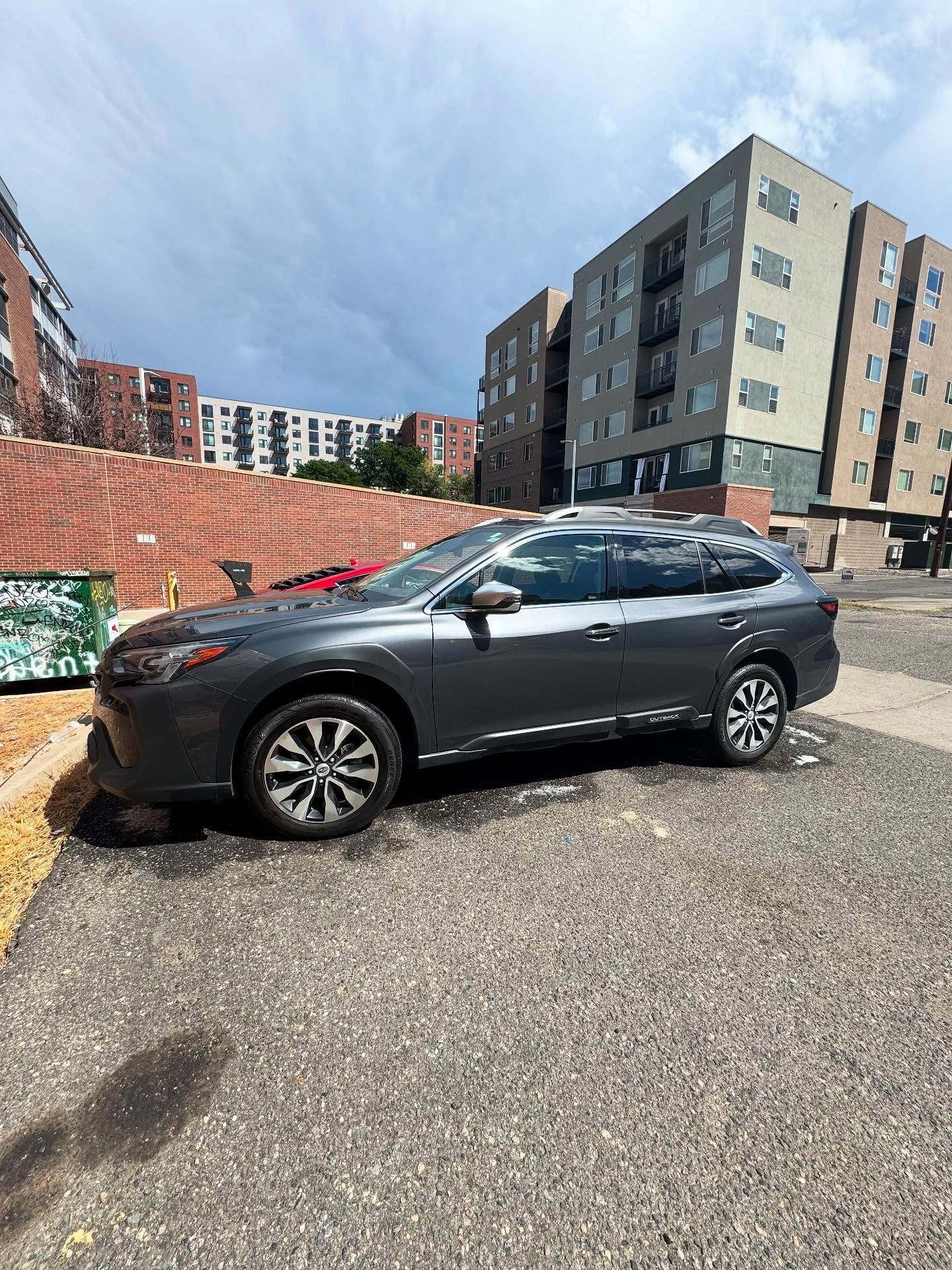 Gray Subaru Outback parked near brick apartment buildings on a sunny day.