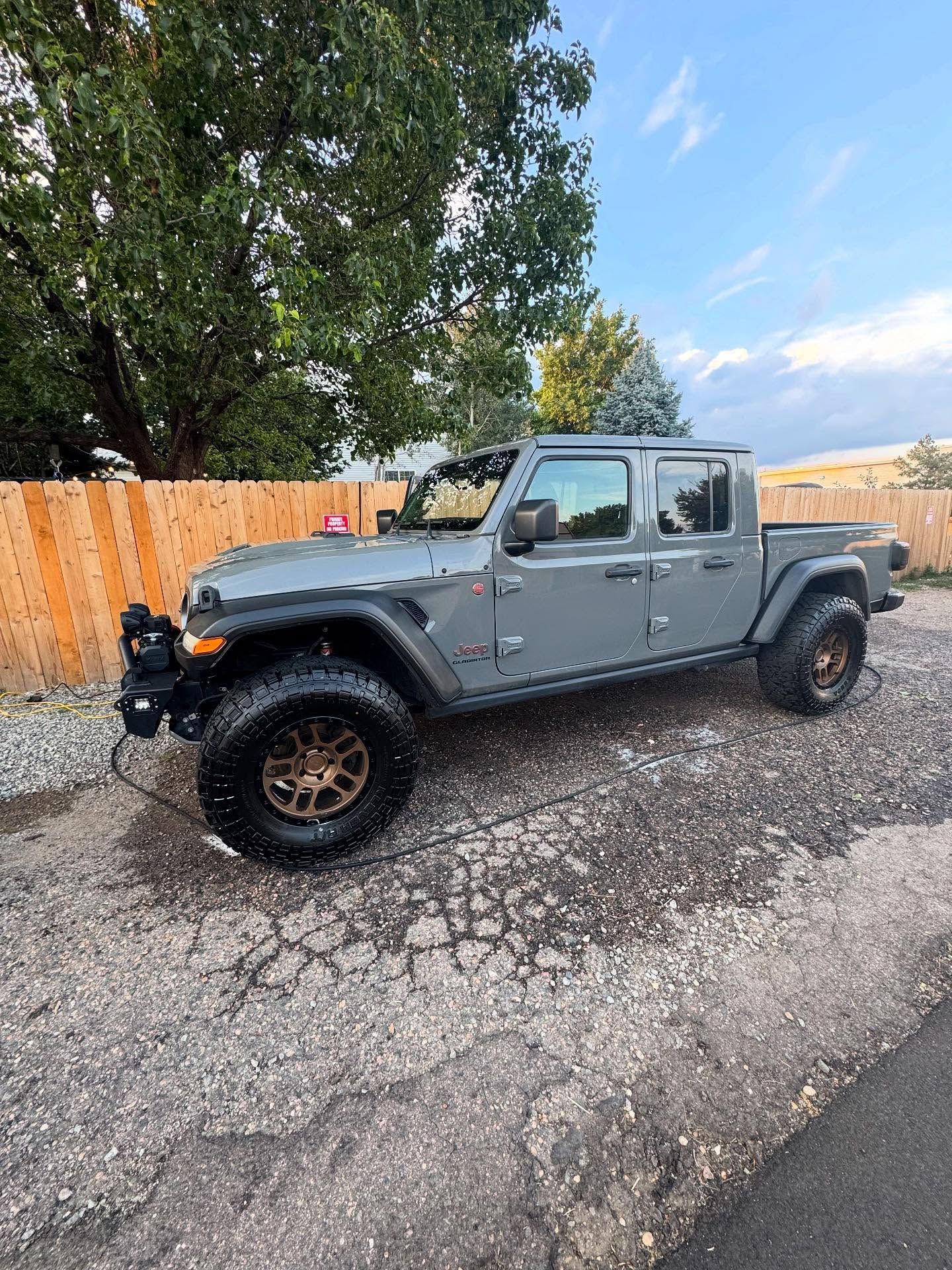 Gray Jeep Gladiator truck parked on cracked pavement. Dark bronze wheels, a wood fence, and trees in background.