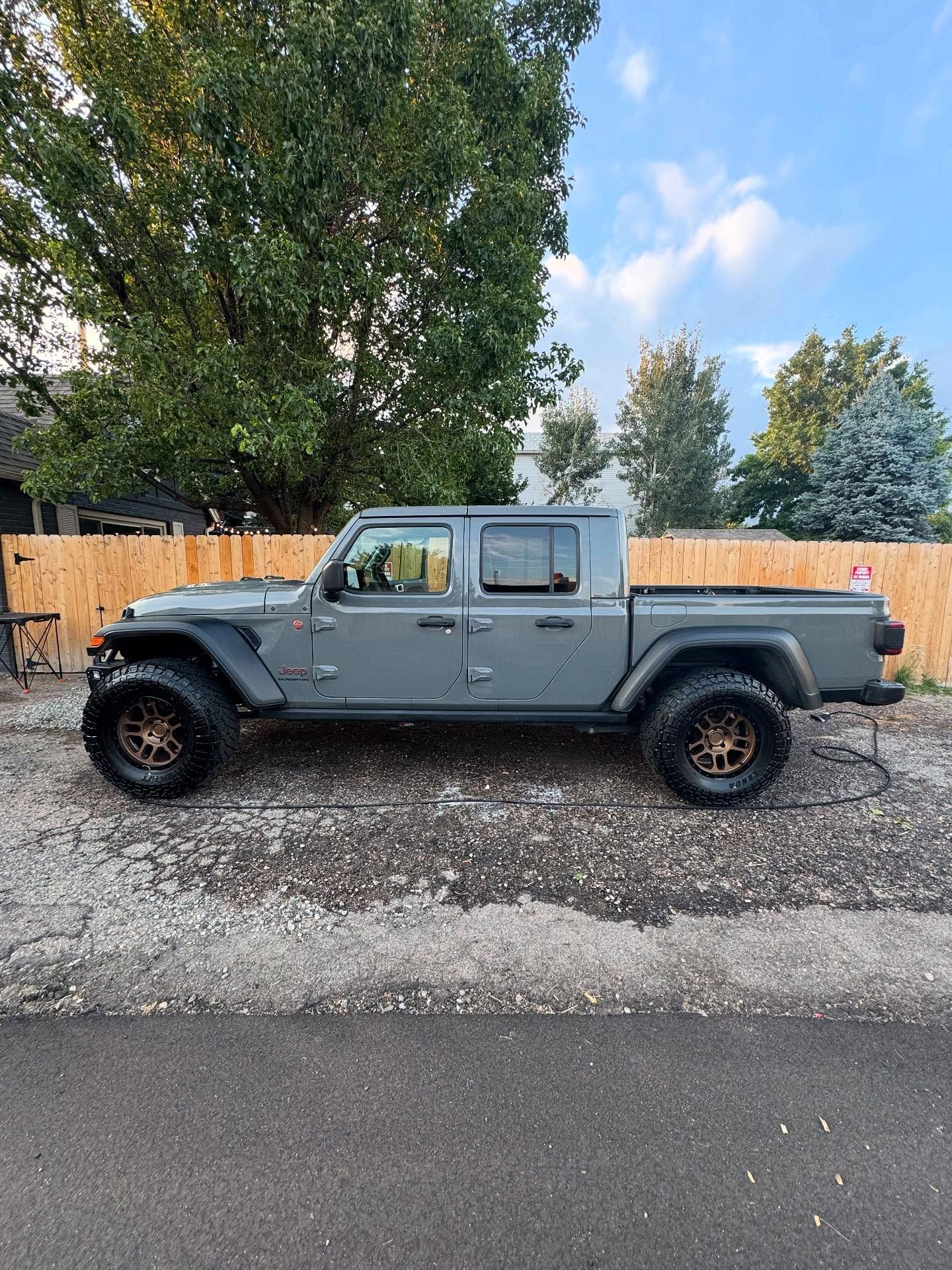 Gray Jeep Gladiator truck with bronze wheels parked on a driveway, surrounded by trees and a wooden fence.