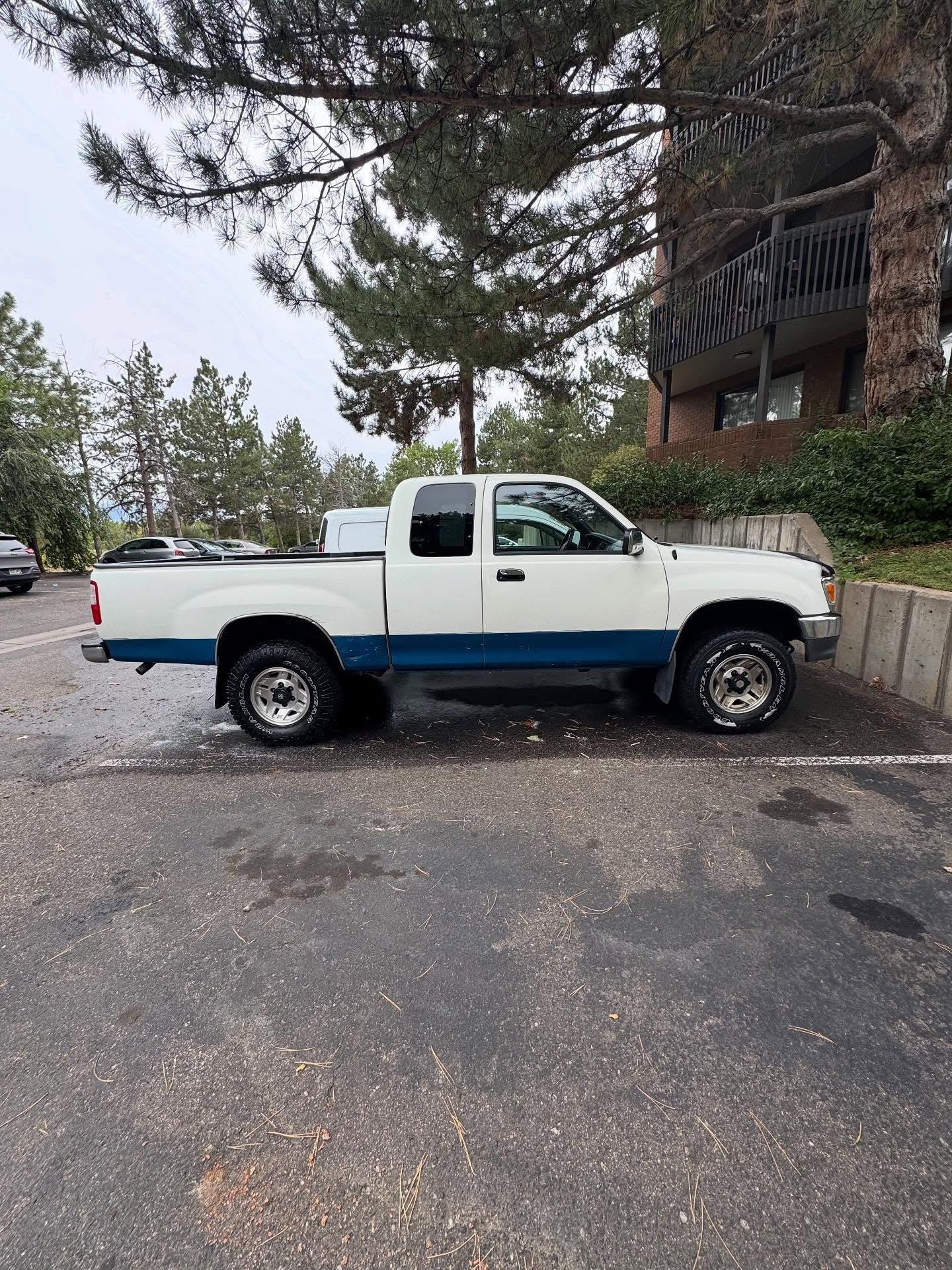 White and blue pickup truck parked on asphalt, near trees and a building.