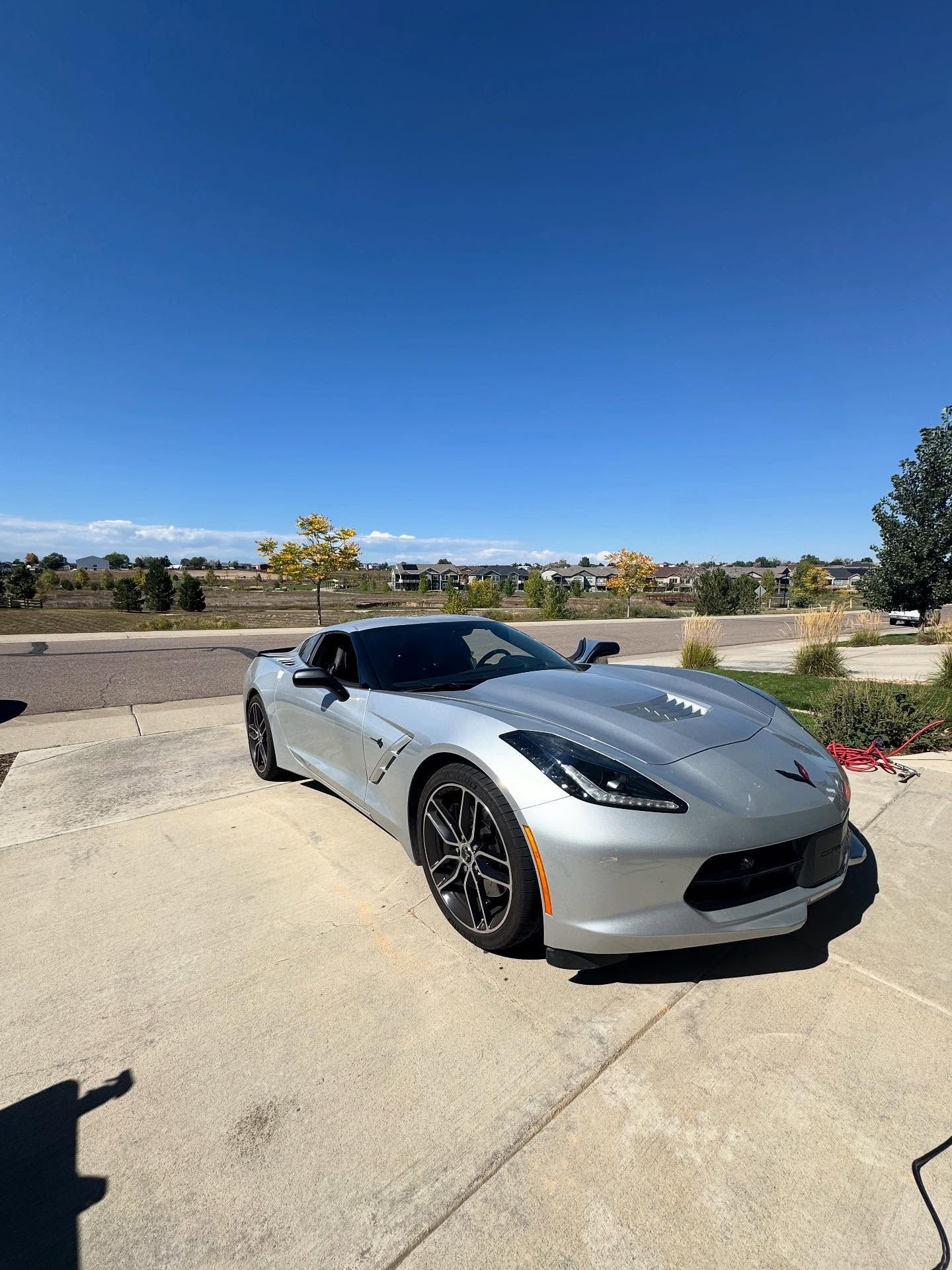 Silver Chevrolet Corvette parked on a concrete driveway, under a clear blue sky.