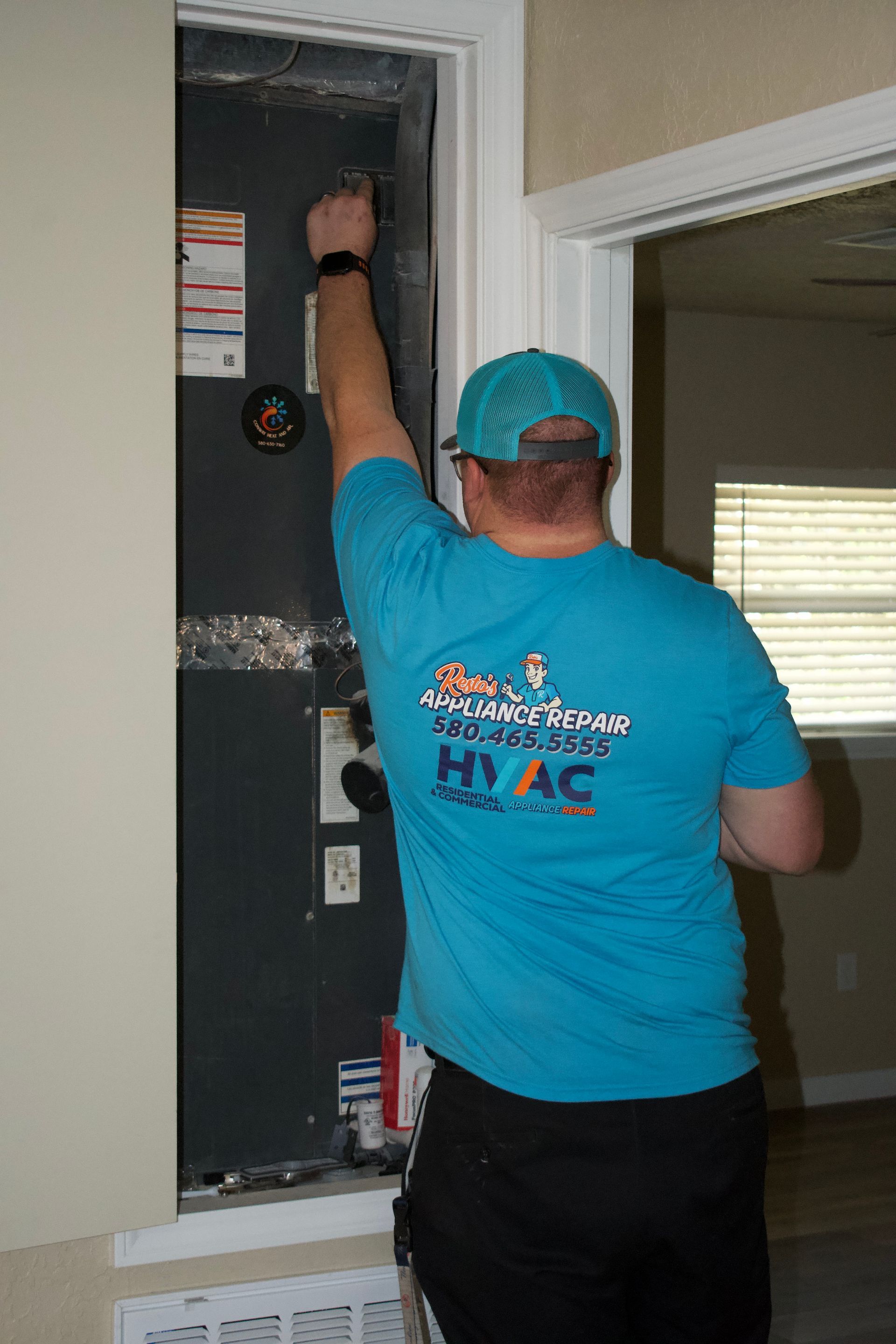 Technician in blue shirt and cap adjusts HVAC unit in a wall opening.