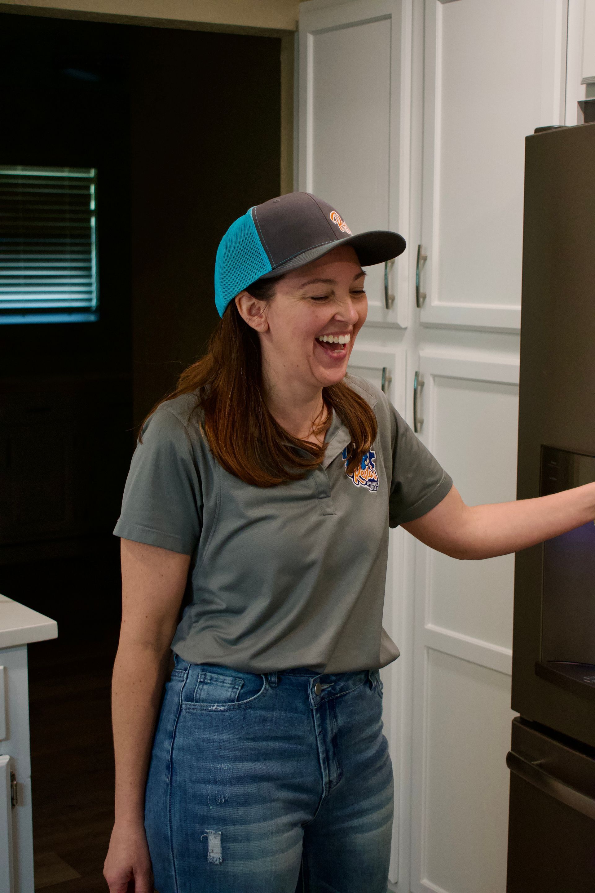 Smiling person in a gray shirt and blue cap standing in a kitchen beside a black refrigerator
