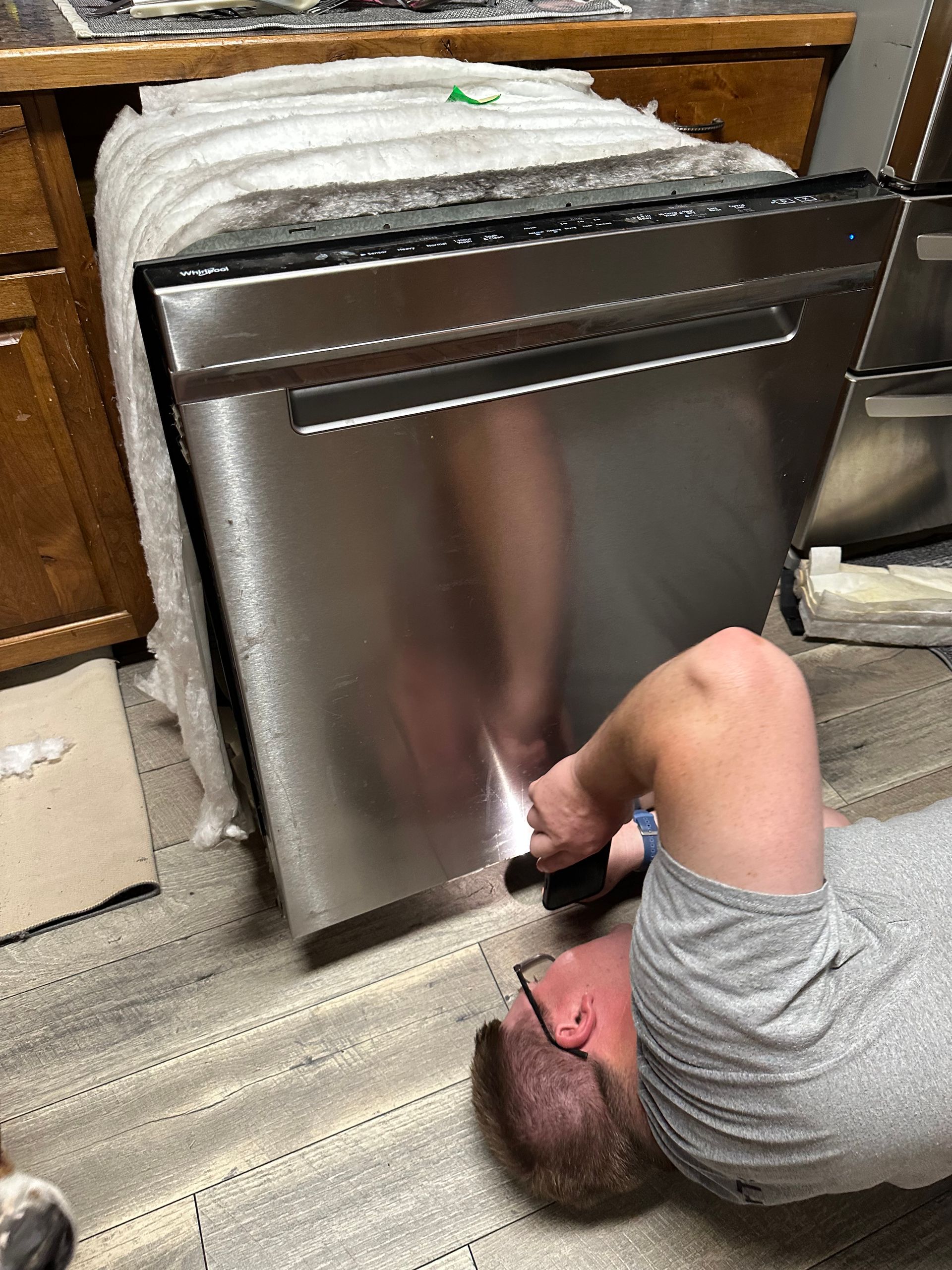 Person lying on floor using a tool to repair a stainless steel dishwasher under a kitchen counter