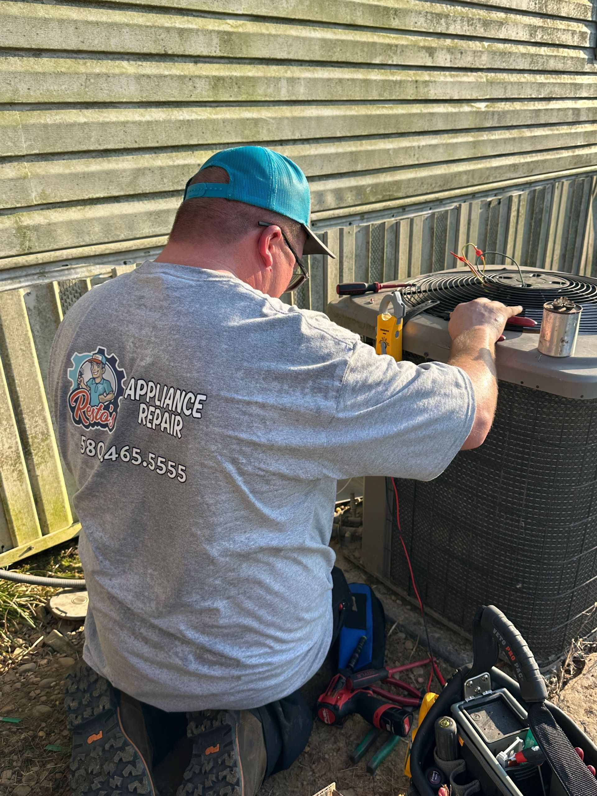 Worker in blue cap servicing outdoor HVAC unit beside a house and tools on the ground