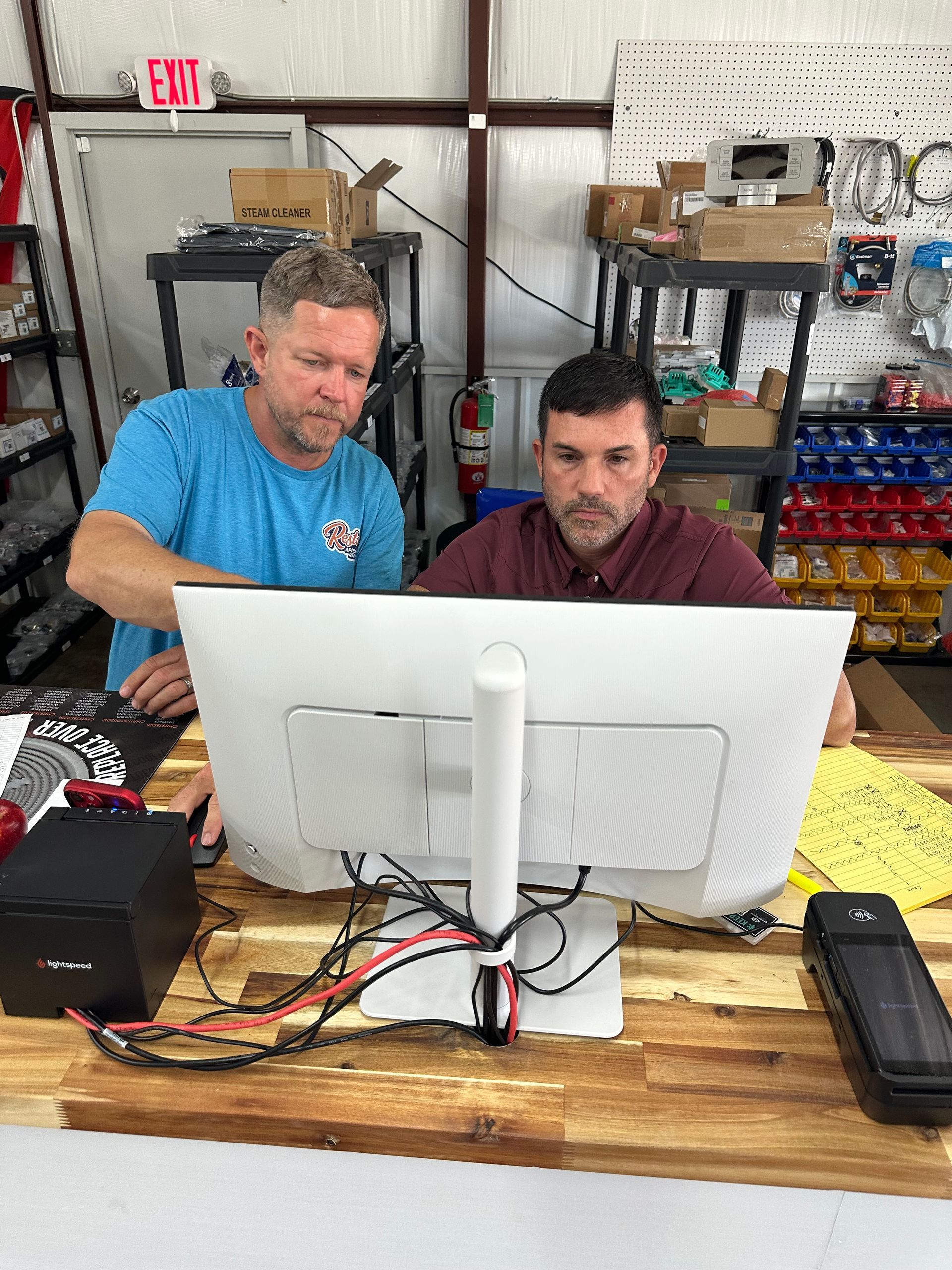 Two men working at a computer desk in a workshop, with tools and supplies on shelves behind them.