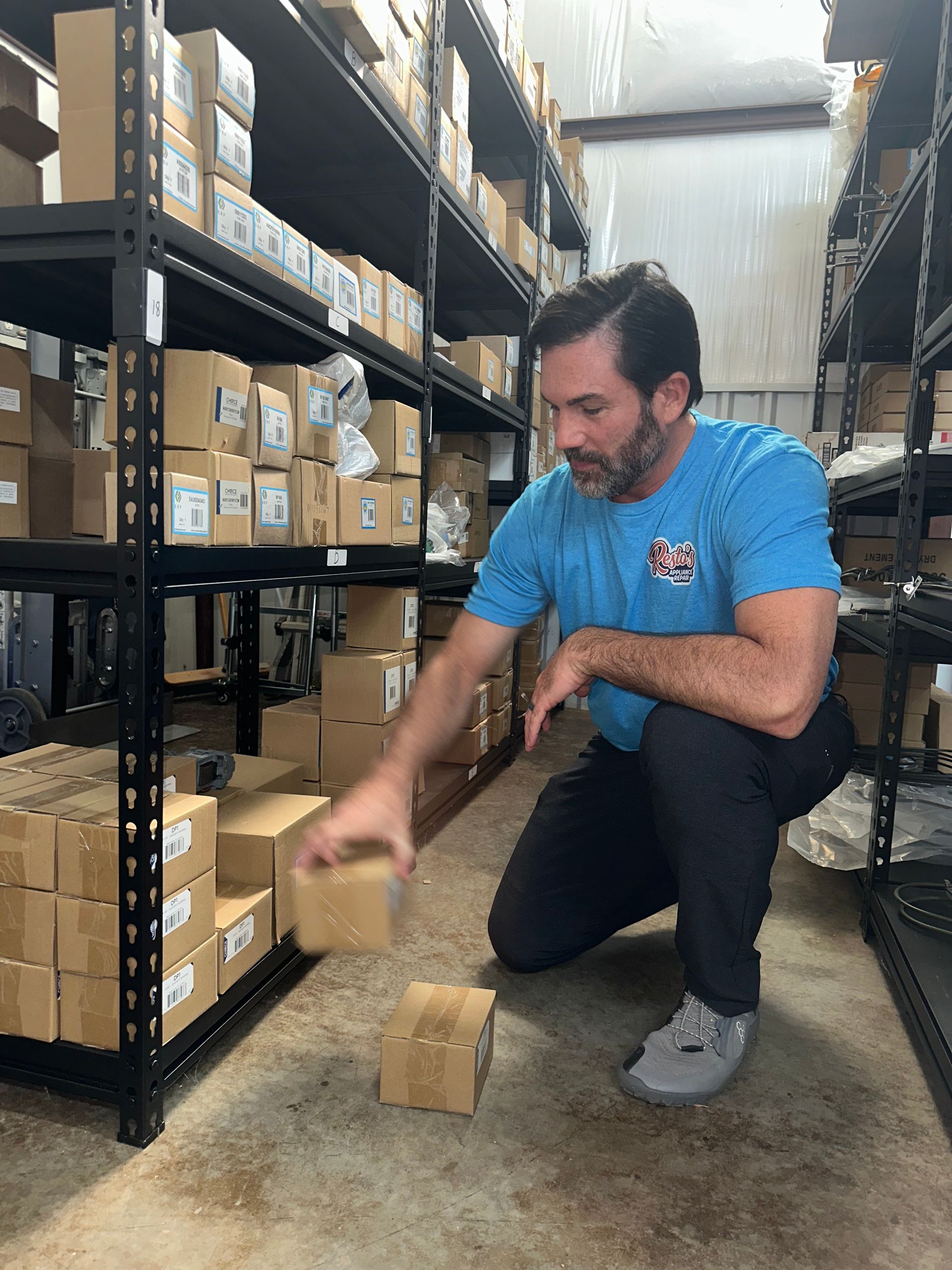 Man crouching in a warehouse aisle, stacking small cardboard boxes near shelves of packaged inventory