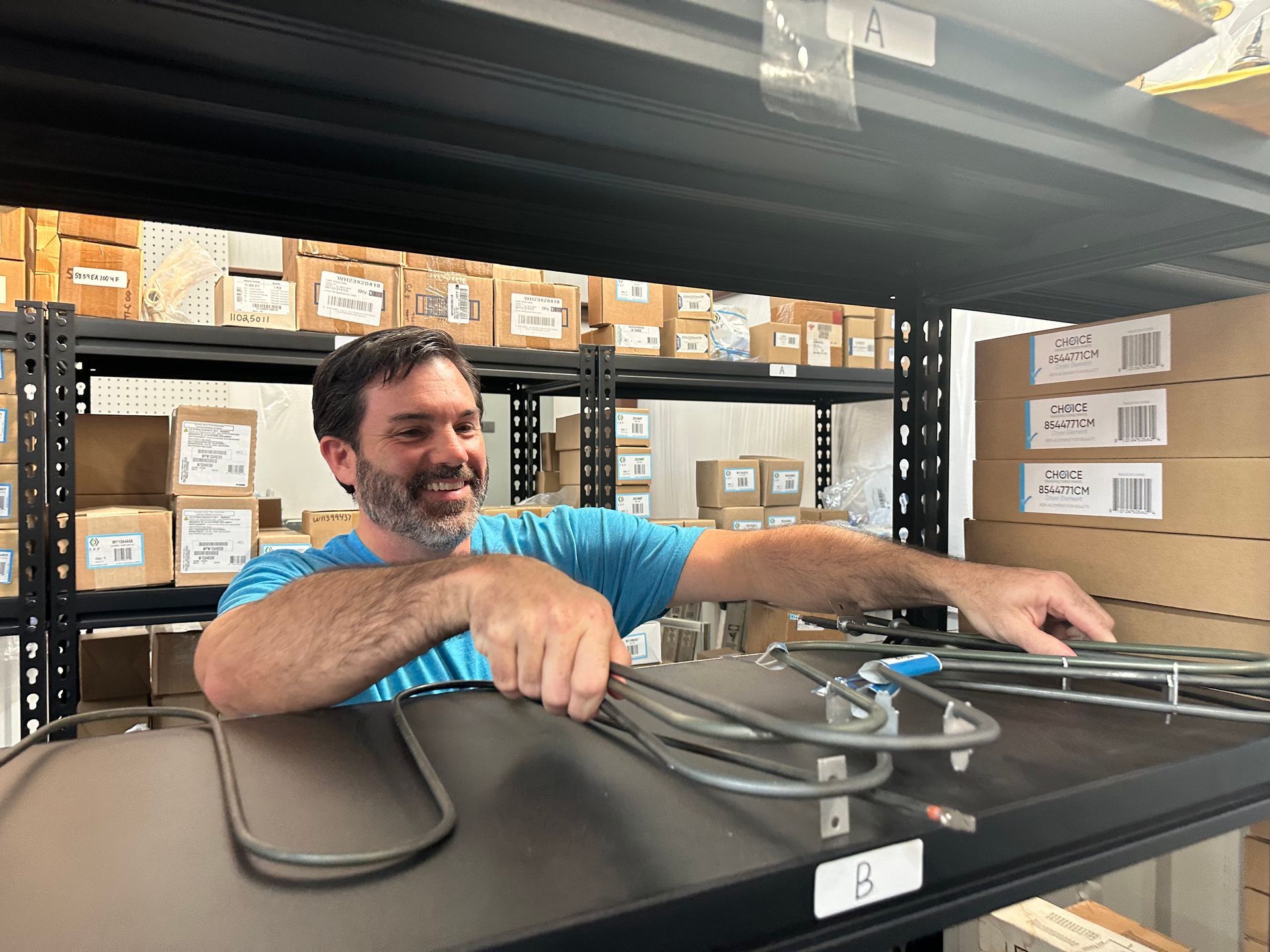 Worker sorting cables on a warehouse shelf, smiling beside stacked boxes and black equipment.