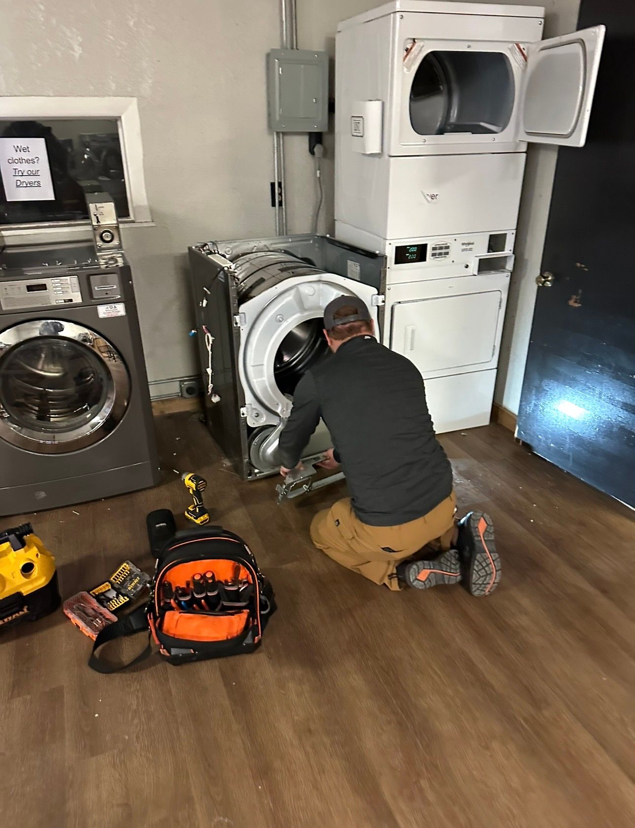 Technician kneeling beside a front-loading washer in a laundry room, with stacked dryer and tools on the floor.