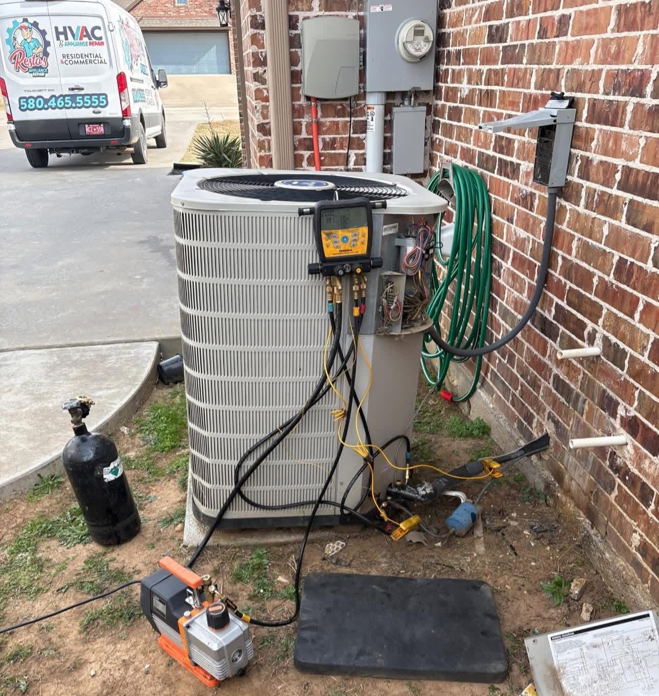 Air conditioner unit with tools and hoses beside a brick wall, service truck in the background