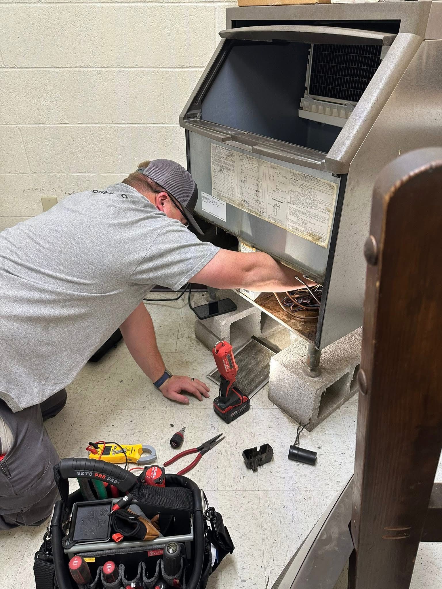Technician kneeling beside a metal fireplace, using tools to repair or clean it, with a toolbox on the floor.
